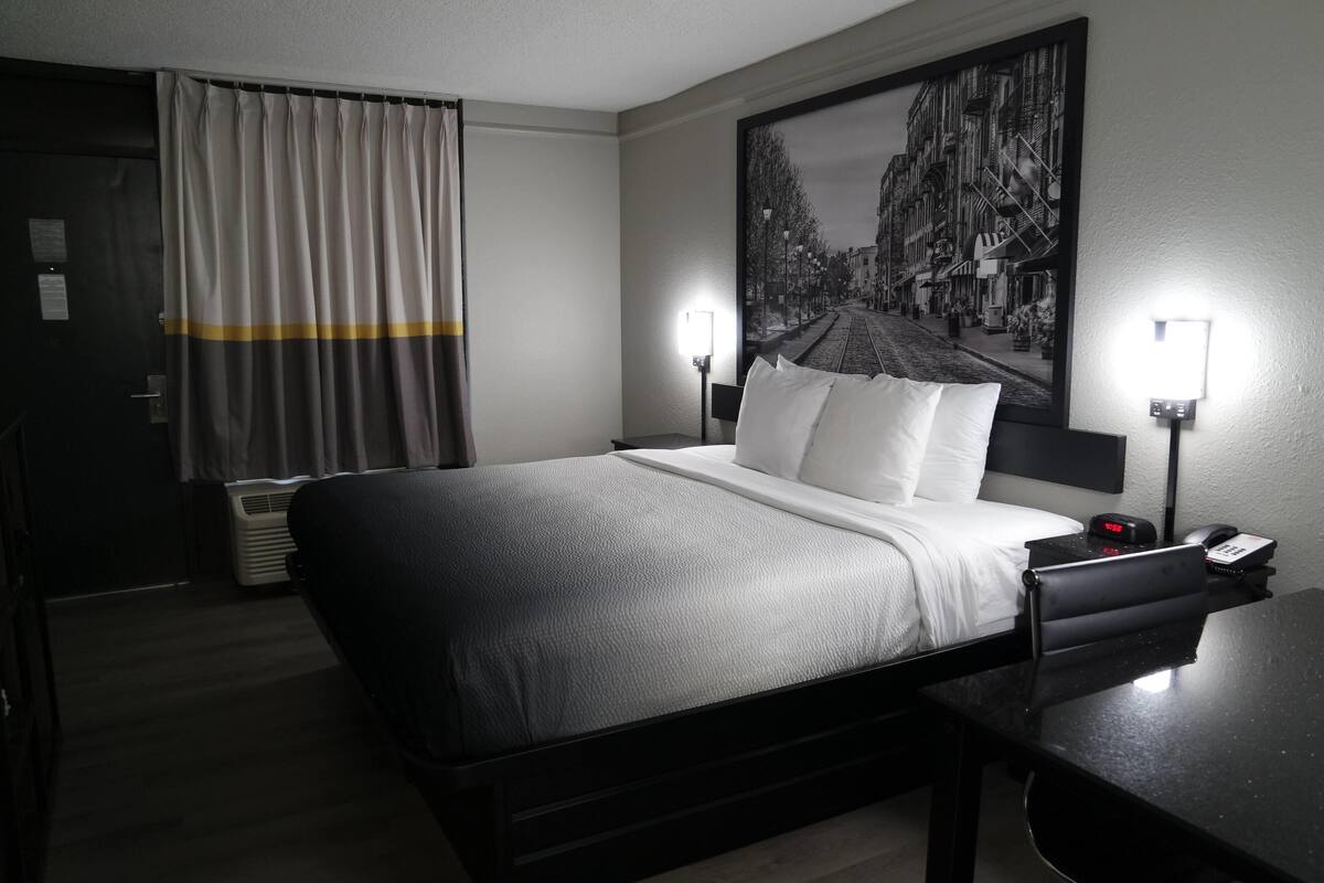 A hotel room with a bed, bedside lamps, a desk, and a large cityscape painting on the wall.