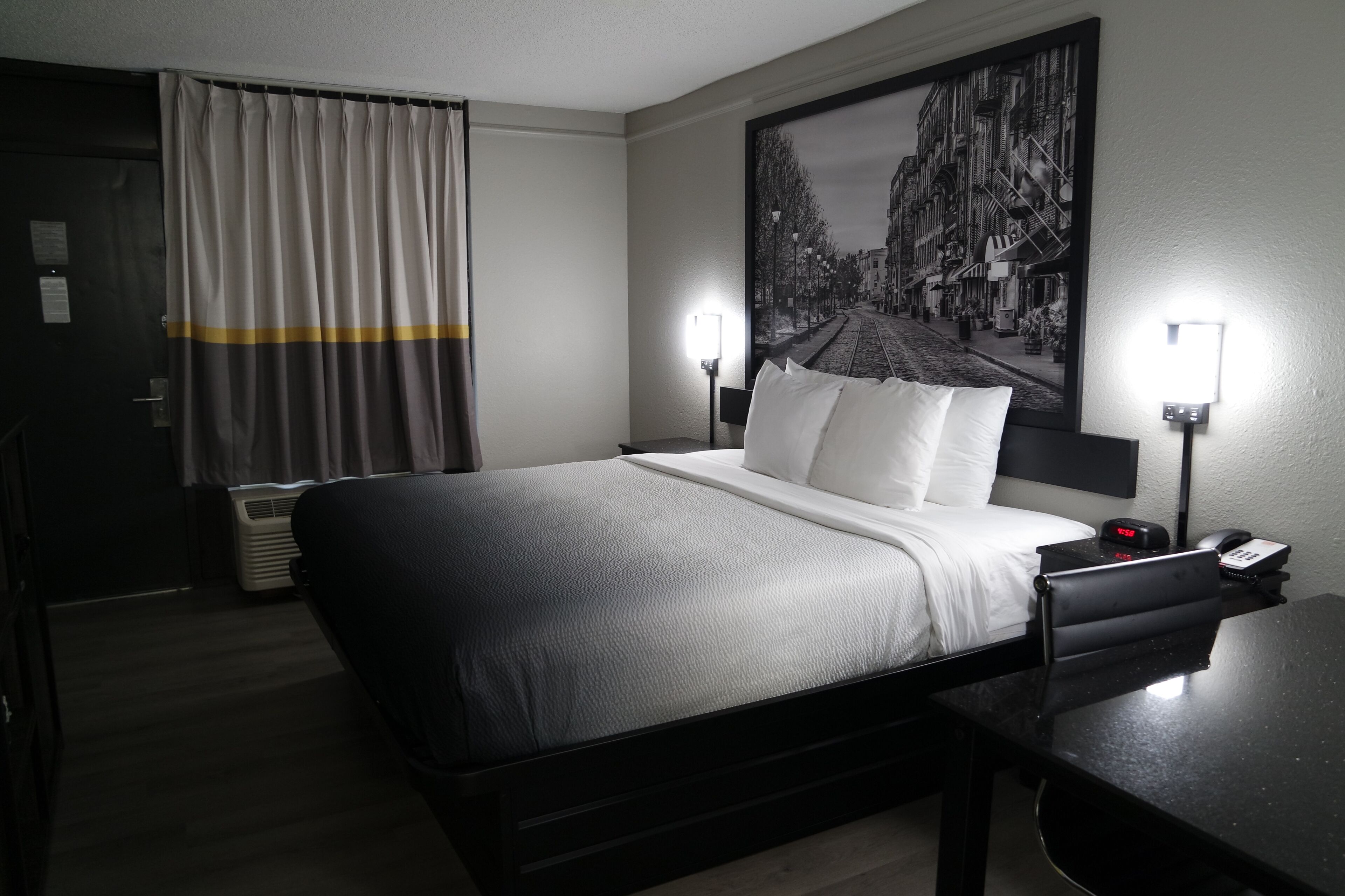 A hotel room with a bed, bedside lamps, a nightstand, a desk, a chair, a window with curtains, and a wall-mounted artwork.