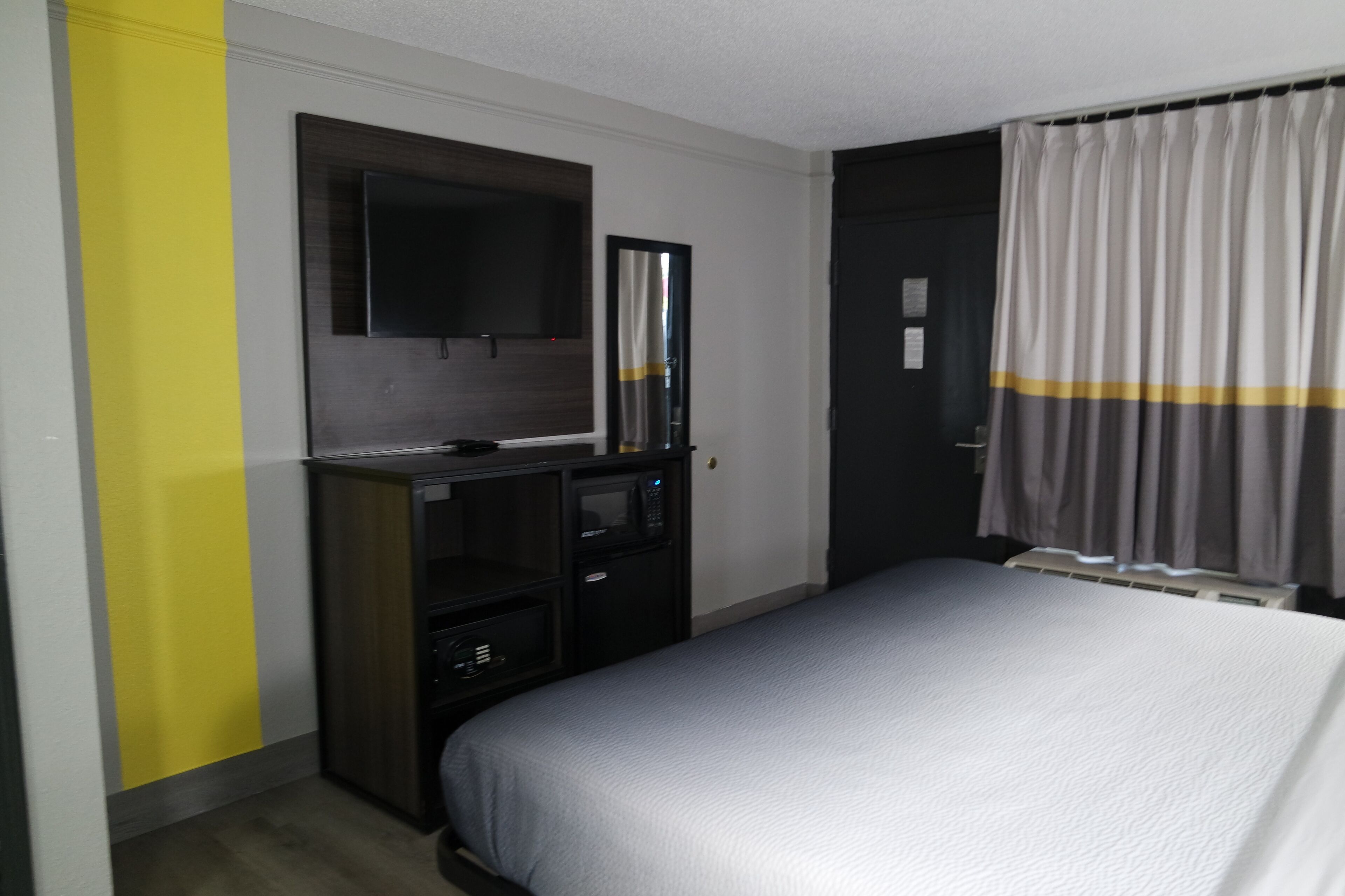 A hotel room with a bed, a desk with a television, a mirror, and a door with curtains.