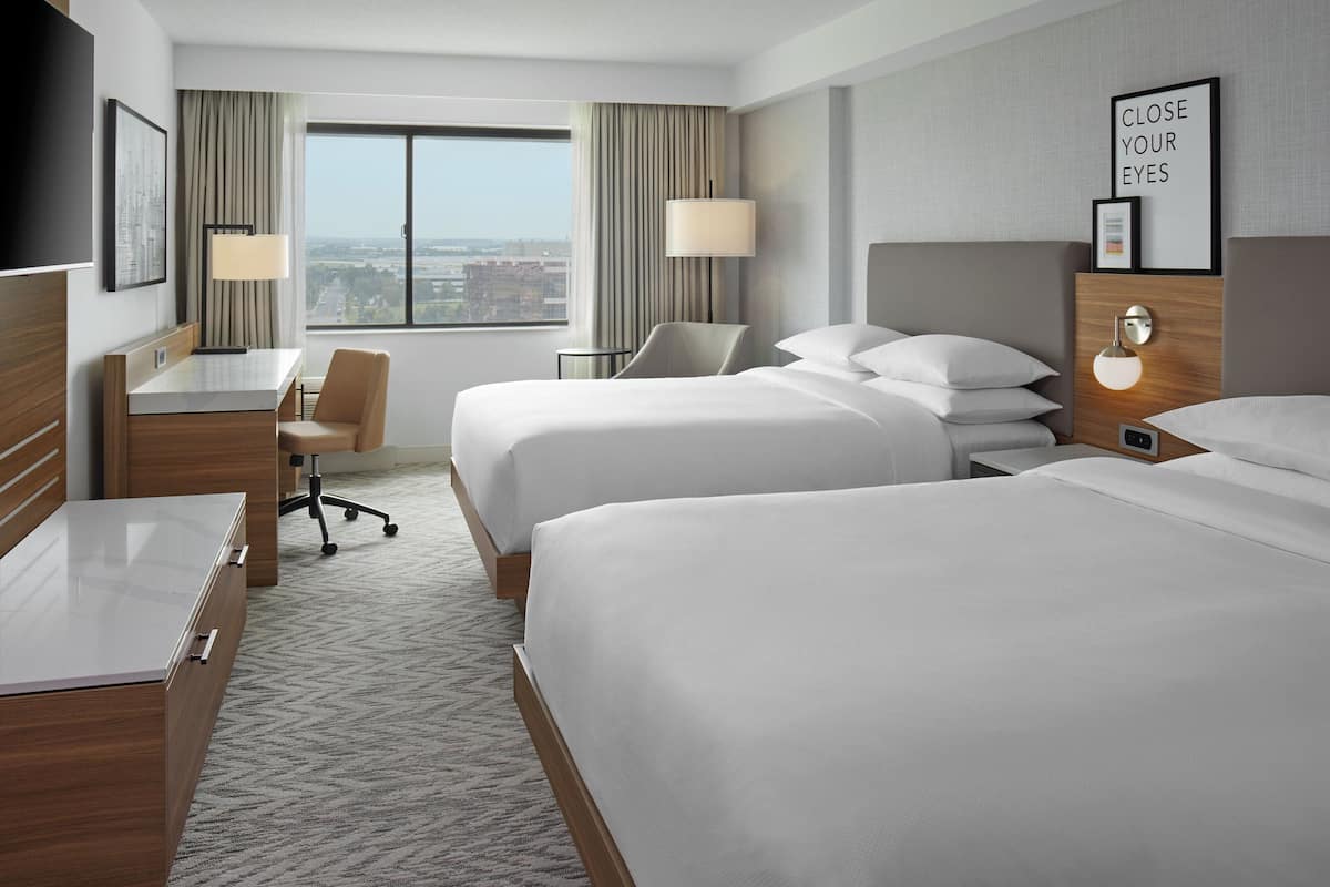 A hotel room with two beds, a desk, a chair, a TV, and a window with a city view.