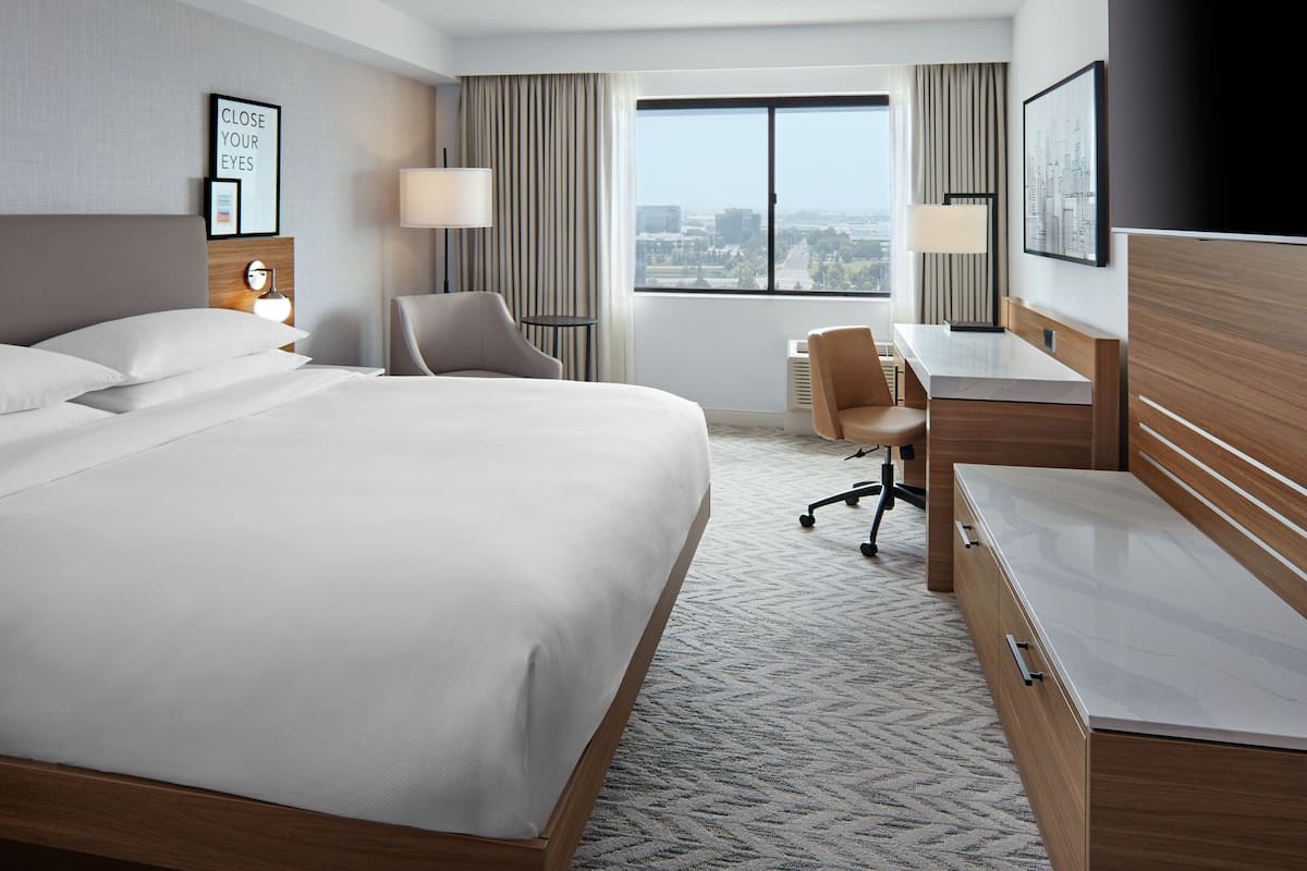 A hotel room with a large bed, a desk, a chair, and a view of the city through the window.