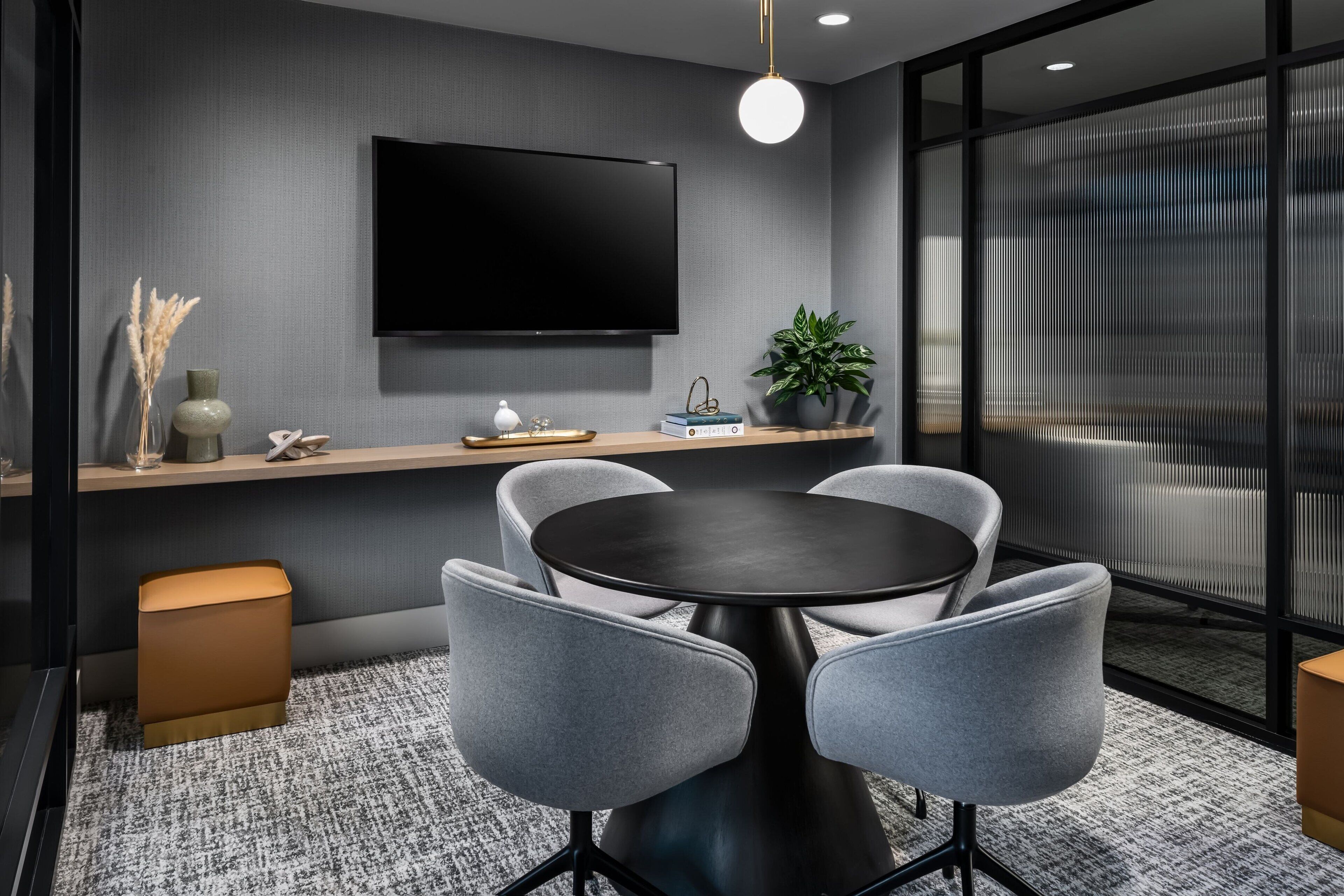 Uma sala de conferências moderna com uma mesa redonda, cadeiras cinzas, uma TV fixada na parede, uma planta e uma estante de madeira com objetos decorativos.