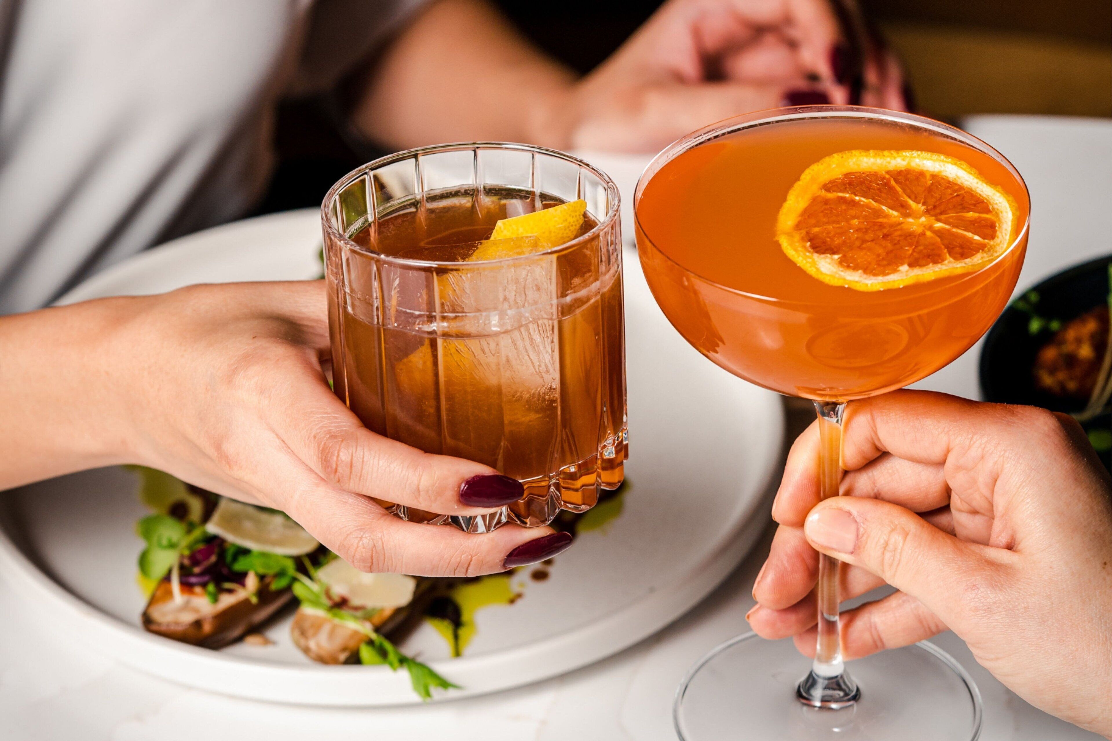 To personer holder hver en drink, den ene i et glas og den anden i et vinglas, med en tallerken mad i baggrunden.