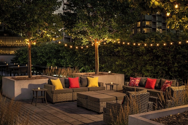 An outdoor seating area with wicker furniture and cushions, surrounded by trees and string lights.