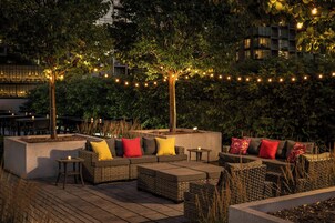 An outdoor seating area with wicker furniture and cushions, surrounded by trees and string lights.