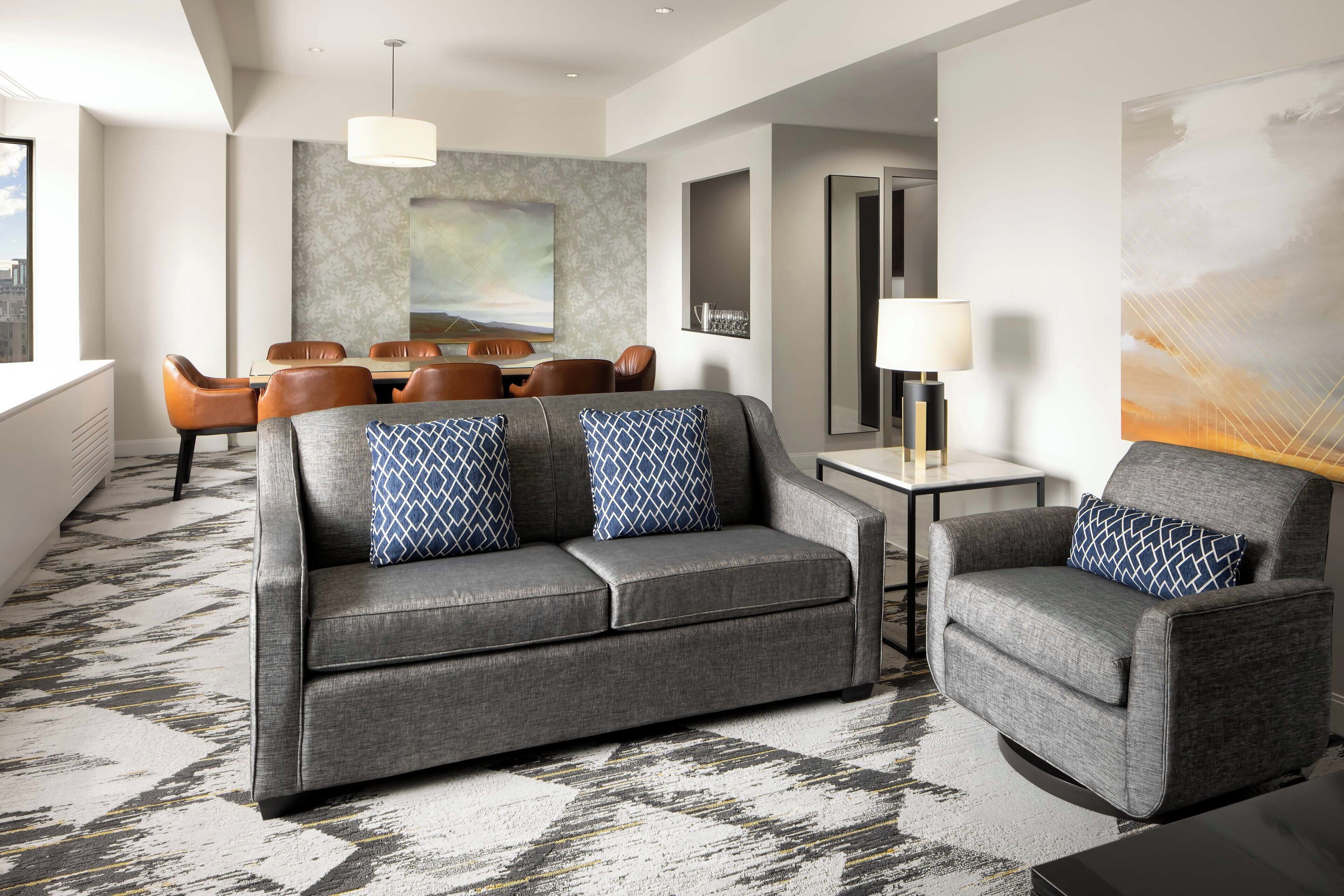 A modern hotel room with a grey sofa, armchair, and matching patterned cushions. A dining area with a table and chairs is visible in the background.