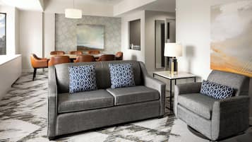 A modern hotel room with a grey sofa, armchair, and matching patterned cushions. A dining area with a table and chairs is visible in the background.