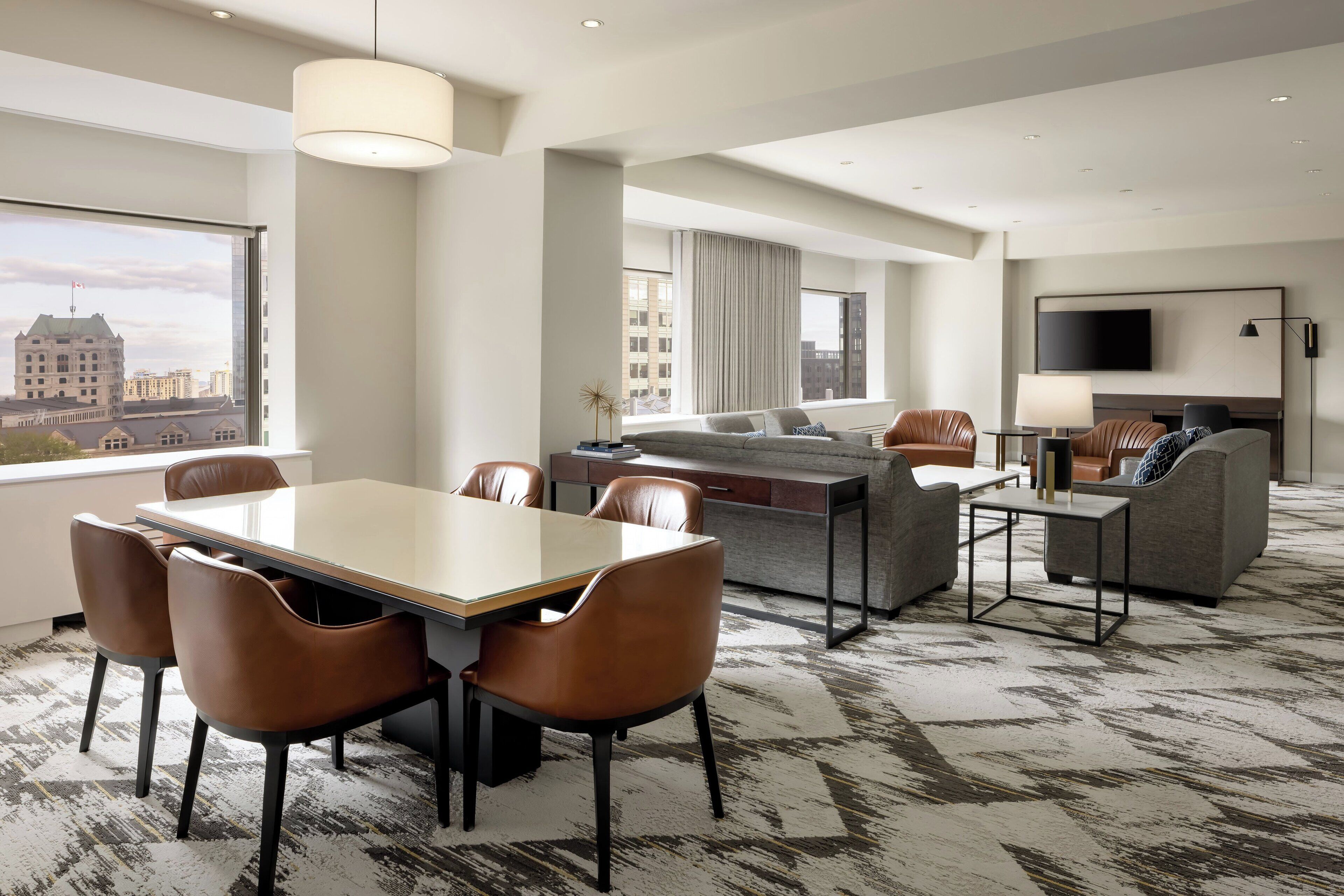 A modern hotel room with a dining area, a sofa set, a flat-screen TV, and a large window offering a city view.