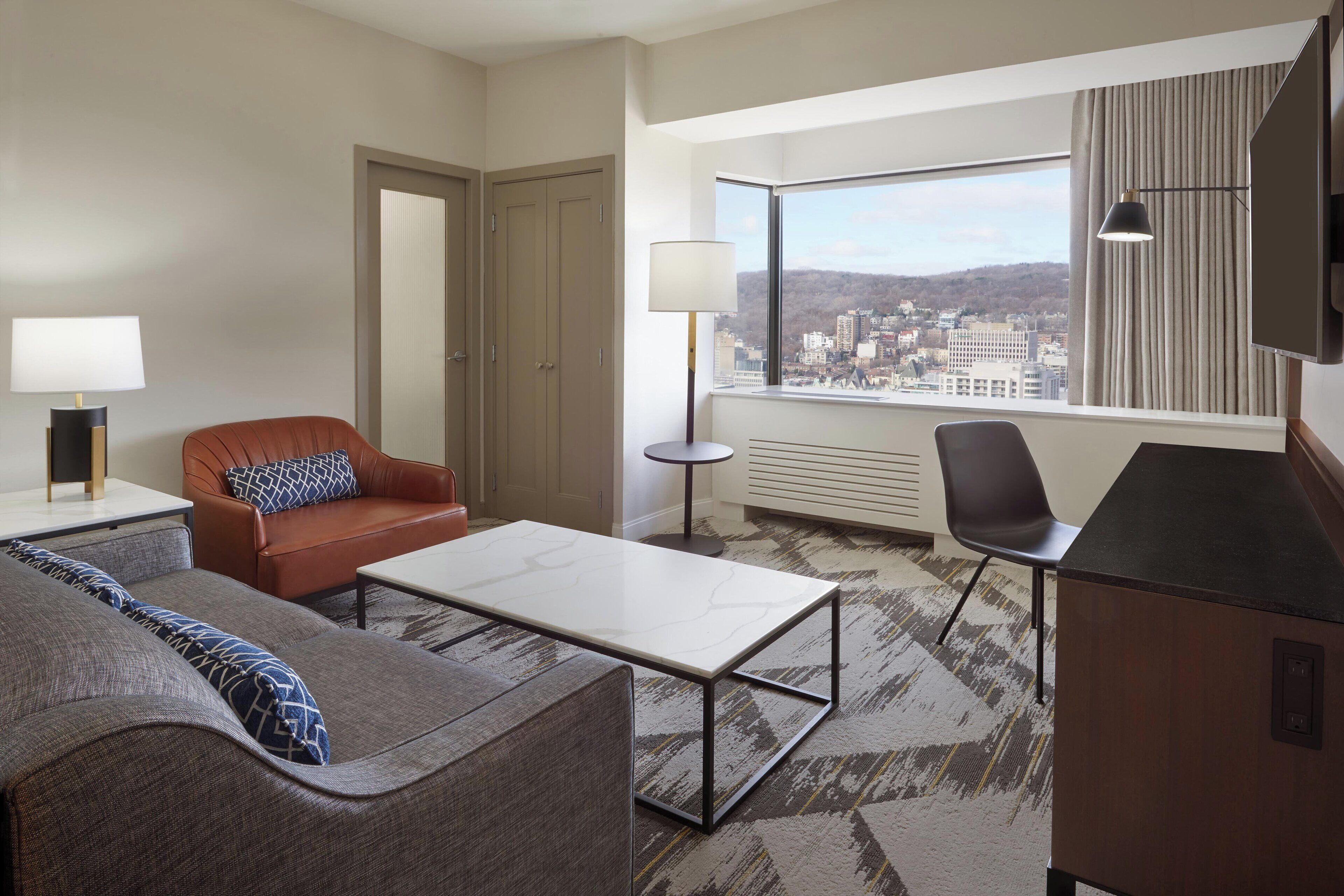 A modern hotel room with a sofa, armchair, and a small table. A city view is visible through the window.