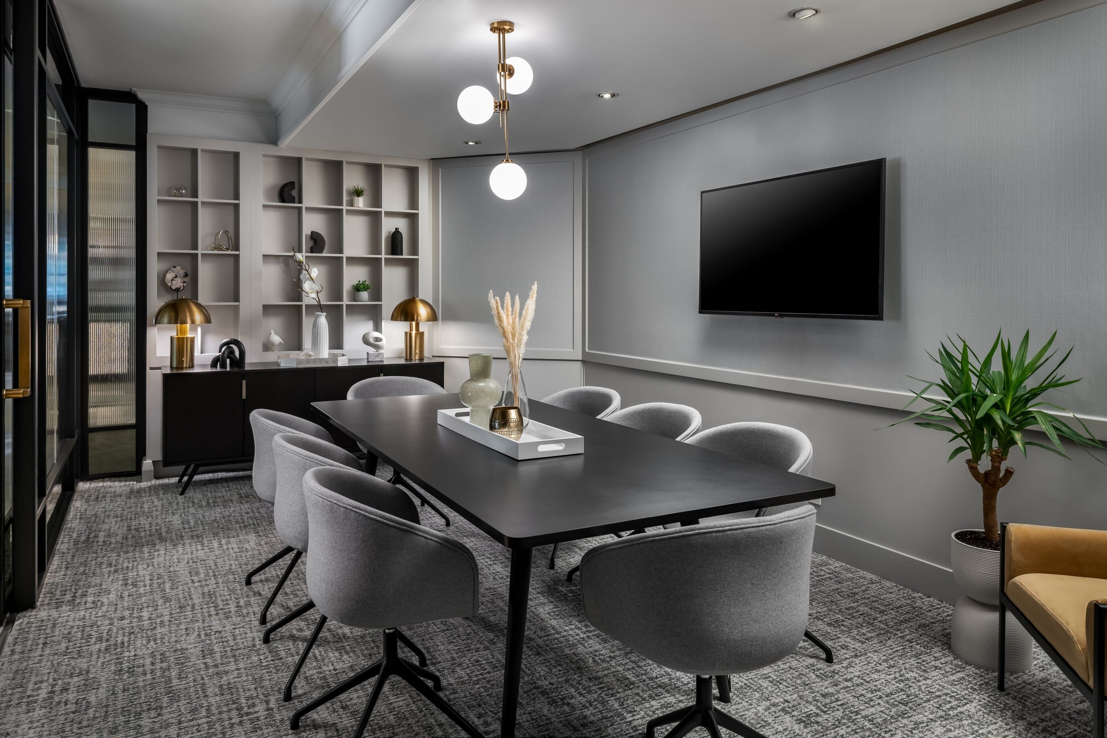 A modern conference room with a long table, six chairs, a flat-screen TV, and a shelving unit with decorative items.