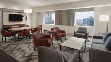 Habitación de hotel con un televisor de pantalla plana grande, zona de comedor con mesa y sillas, sofá, mesita de centro y ventanas amplias con vistas a la ciudad.