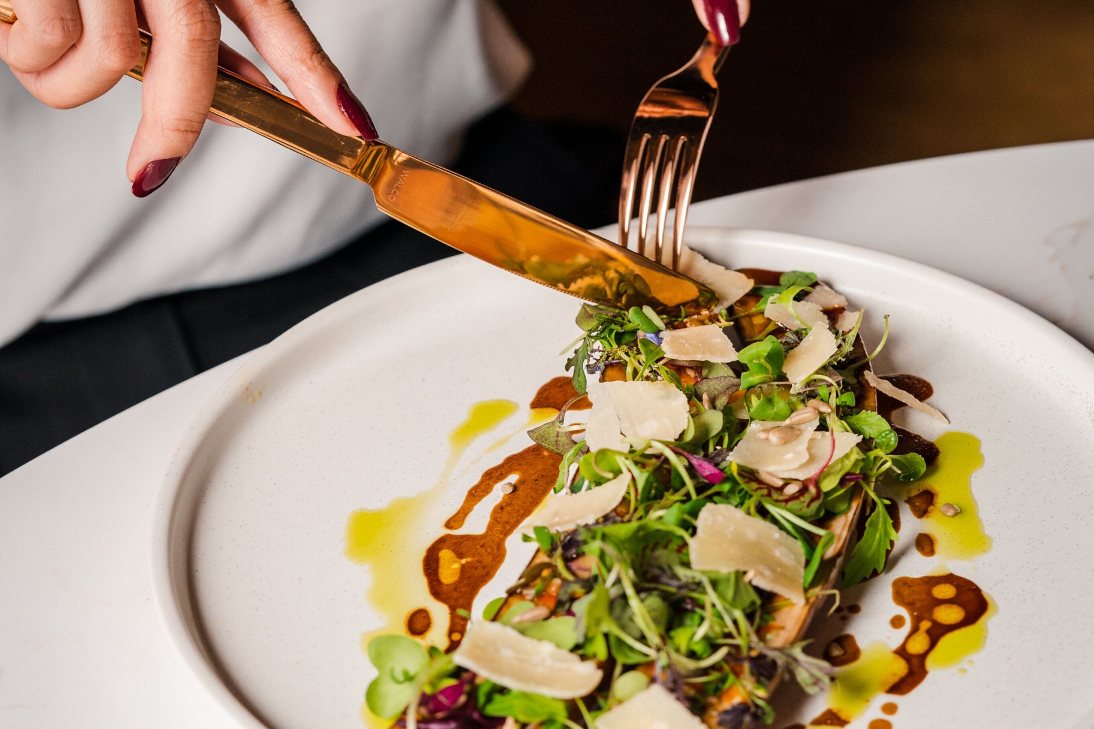 A person using a gold-colored knife and fork to cut a dish with greens and shaved cheese on a white plate.
