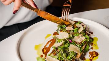 A person using a gold-colored knife and fork to cut a dish with greens and shaved cheese on a white plate.