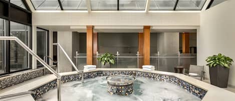 A modern indoor pool area with a glass ceiling, a central hot tub, and a seating area with plants.