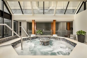 A modern indoor pool area with a glass ceiling, a central hot tub, and a seating area with plants.