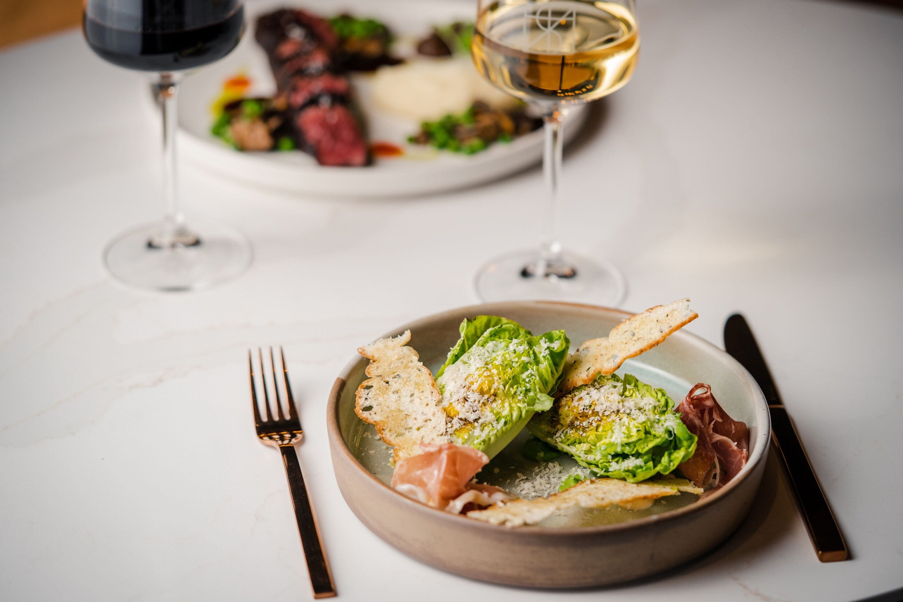 A dining table with a plate of food, a fork, a knife, and two glasses of wine.