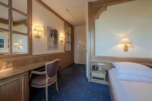 Habitación de hotel con cama, escritorio con silla, mesita de noche pequeña y lámparas fijadas a la pared.