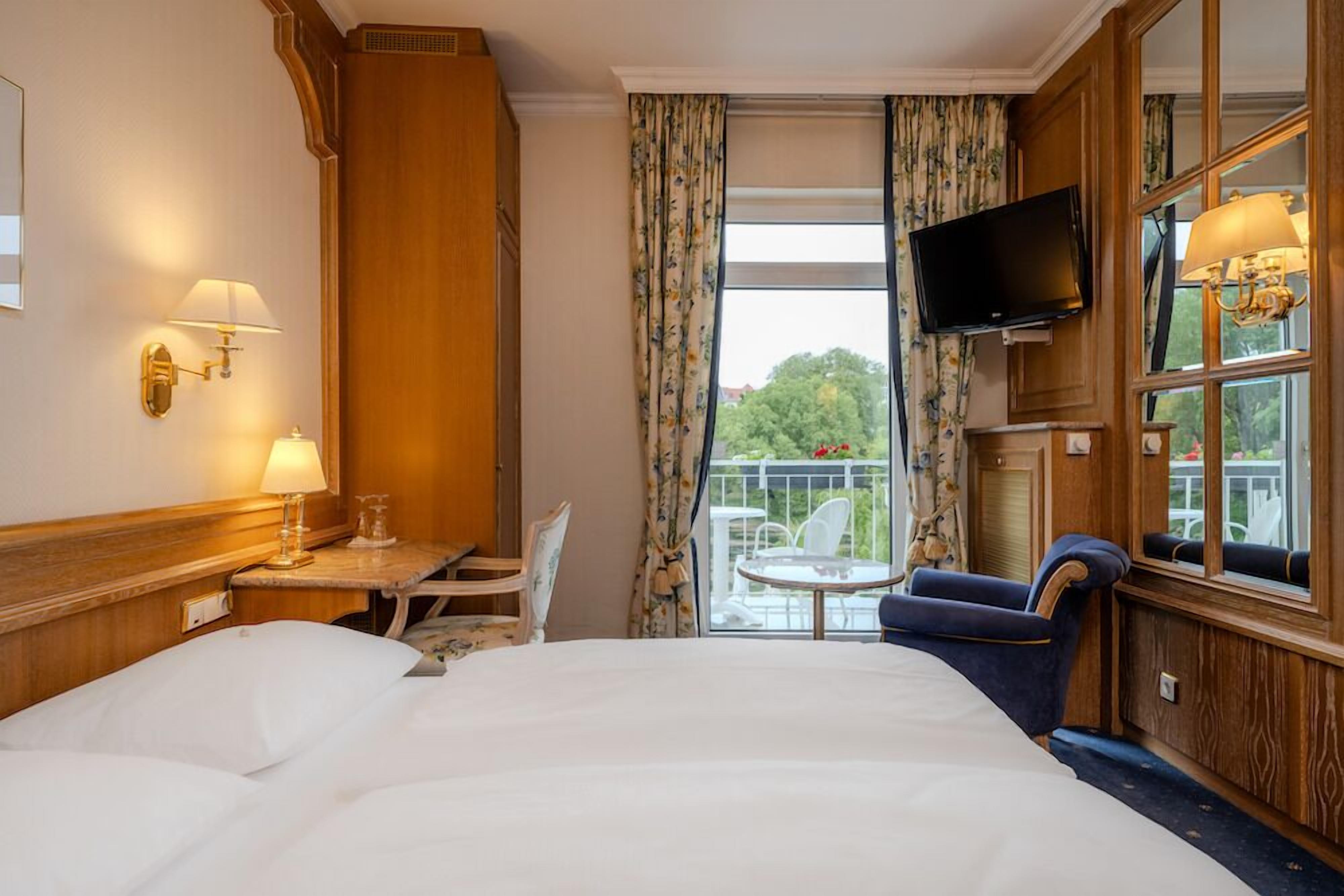 Una habitación de hotel con una cama grande, un escritorio con lámpara, una silla, un televisor montado en la pared y un balcón con mesa y sillas.