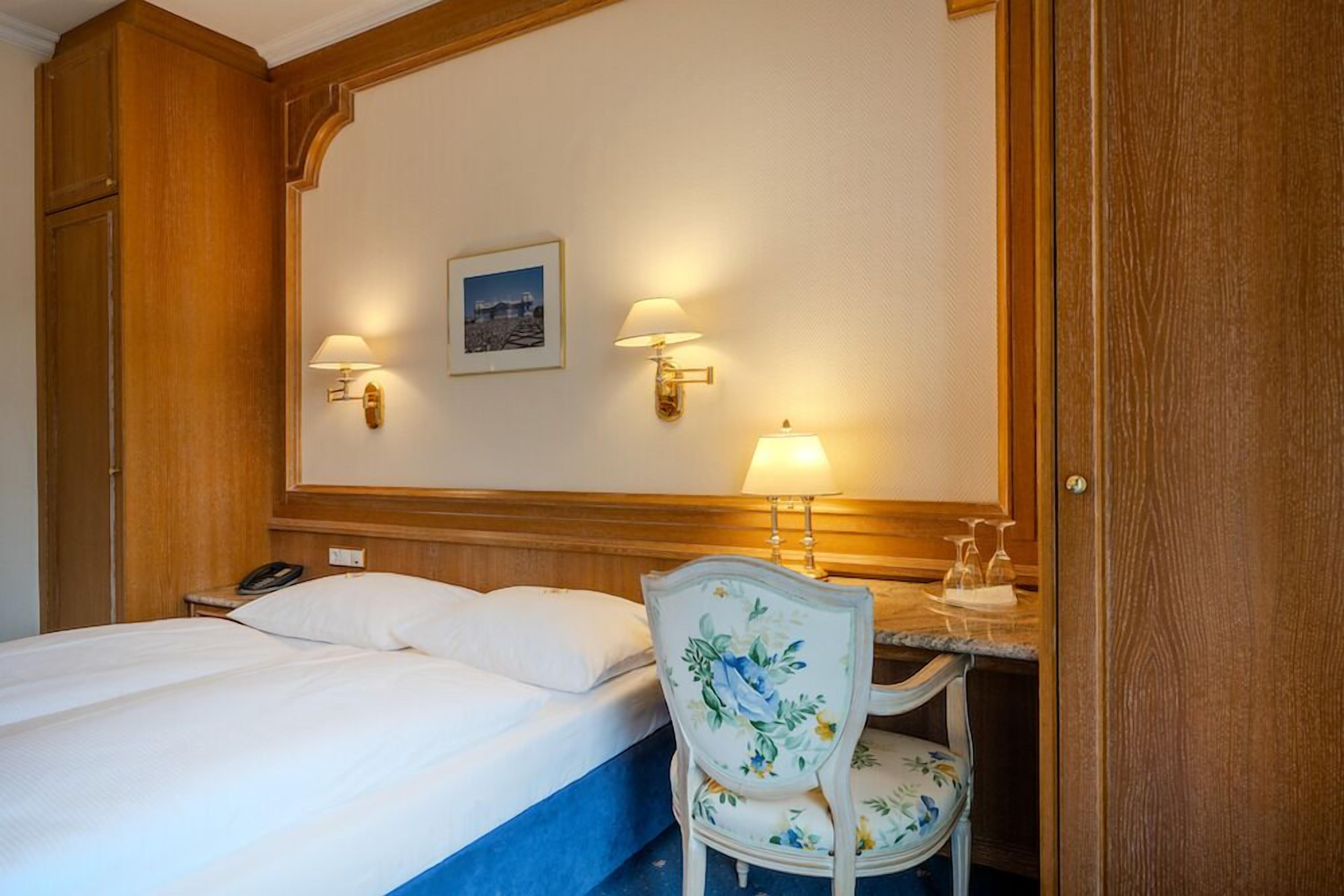 A hotel room with a bed, two wall-mounted lamps, a framed picture, a telephone, and a floral chair.
