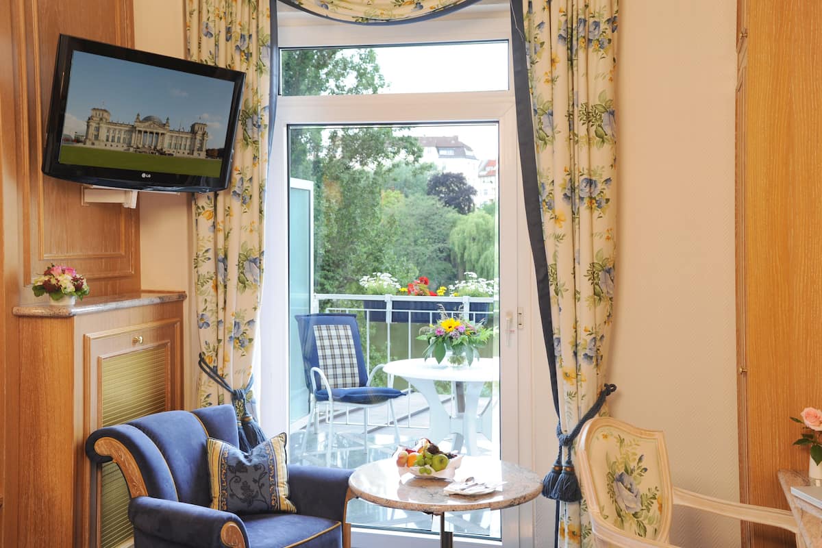 A room with a television mounted on the wall, a sofa, a chair, a small round table with a bowl of fruit, and a balcony with a view of greenery and buildings.
