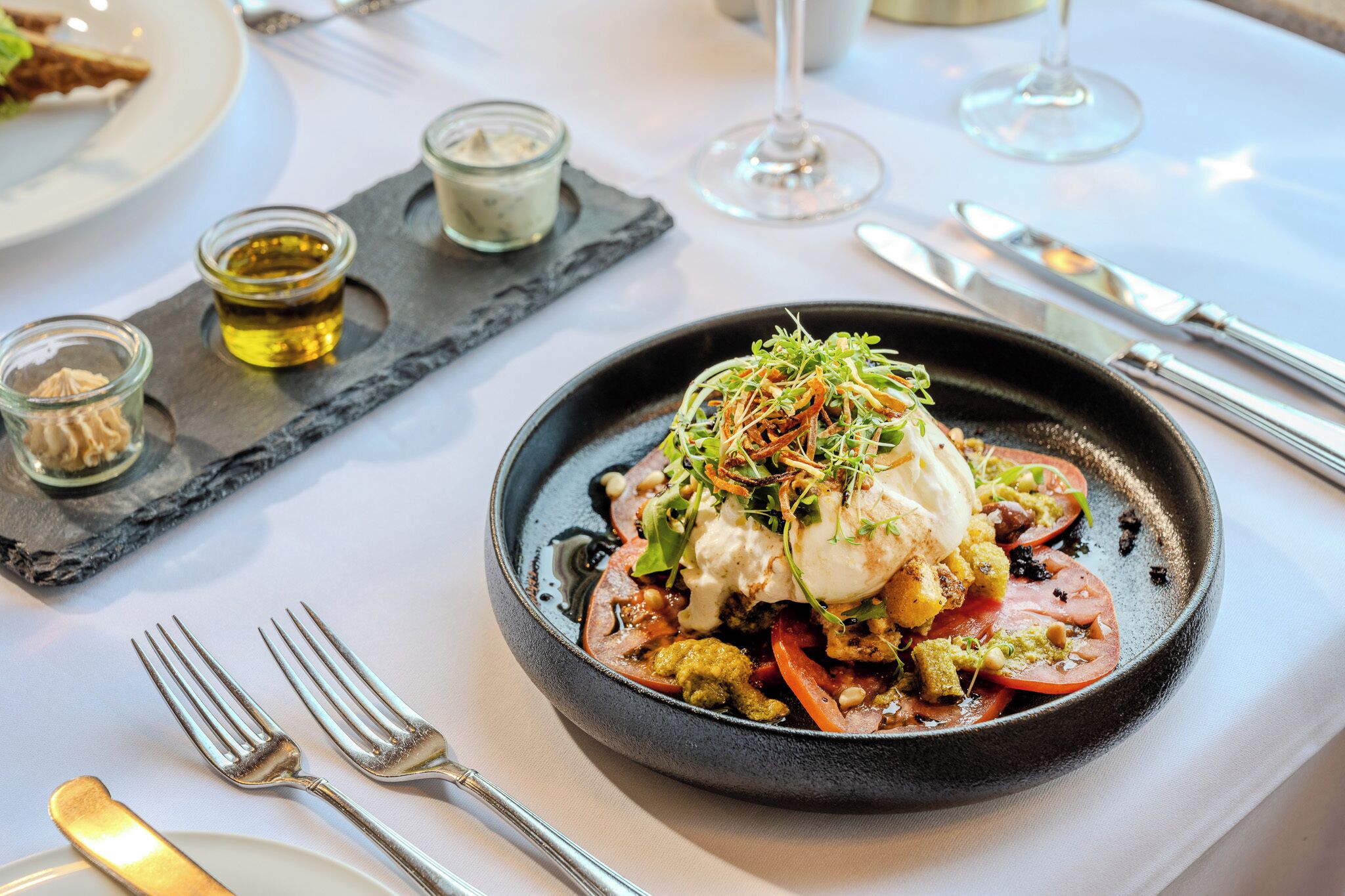 A black plate with a dish of food, including a poached egg, vegetables, and a sauce, on a white tablecloth.