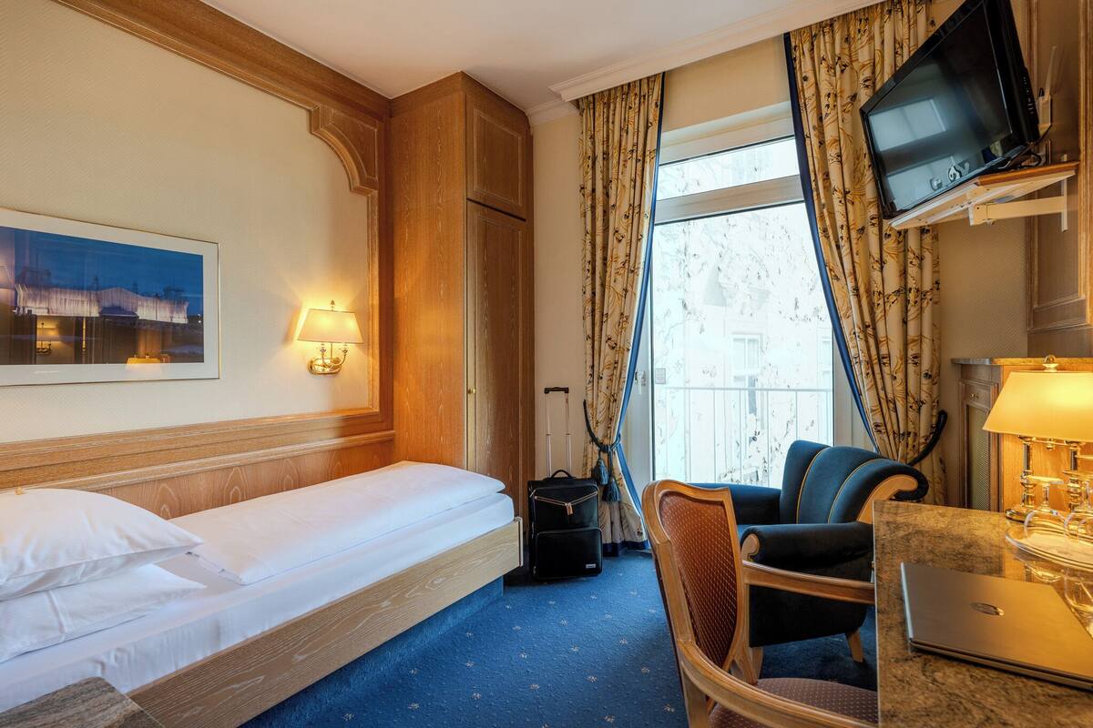 A hotel room with a bed, a desk with a laptop, a chair, a television, and a window with curtains.