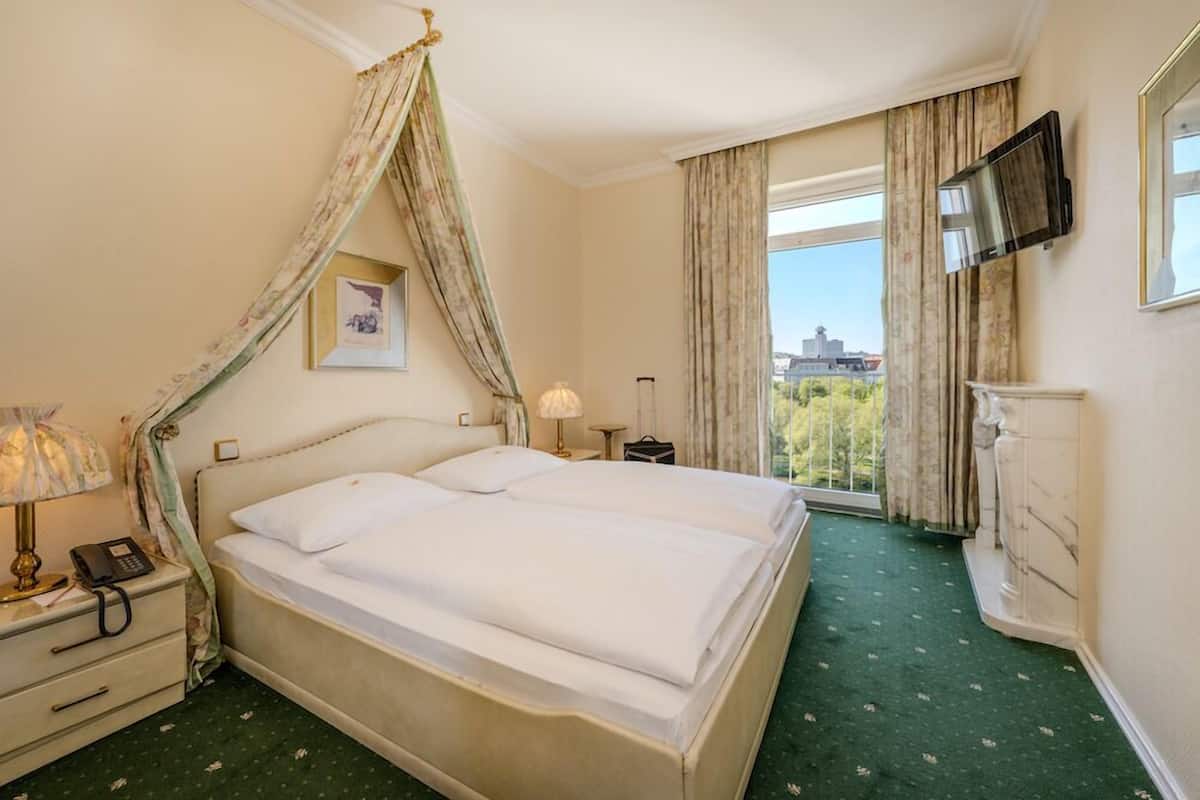 A hotel room with a large bed, a bedside telephone, a television mounted on the wall, and a view of the city through the window.