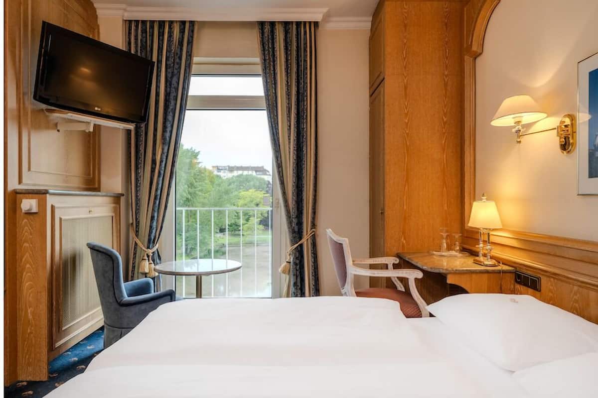 A hotel room with a large bed, a television mounted on the wall, a small round table with a chair, and a balcony with curtains.