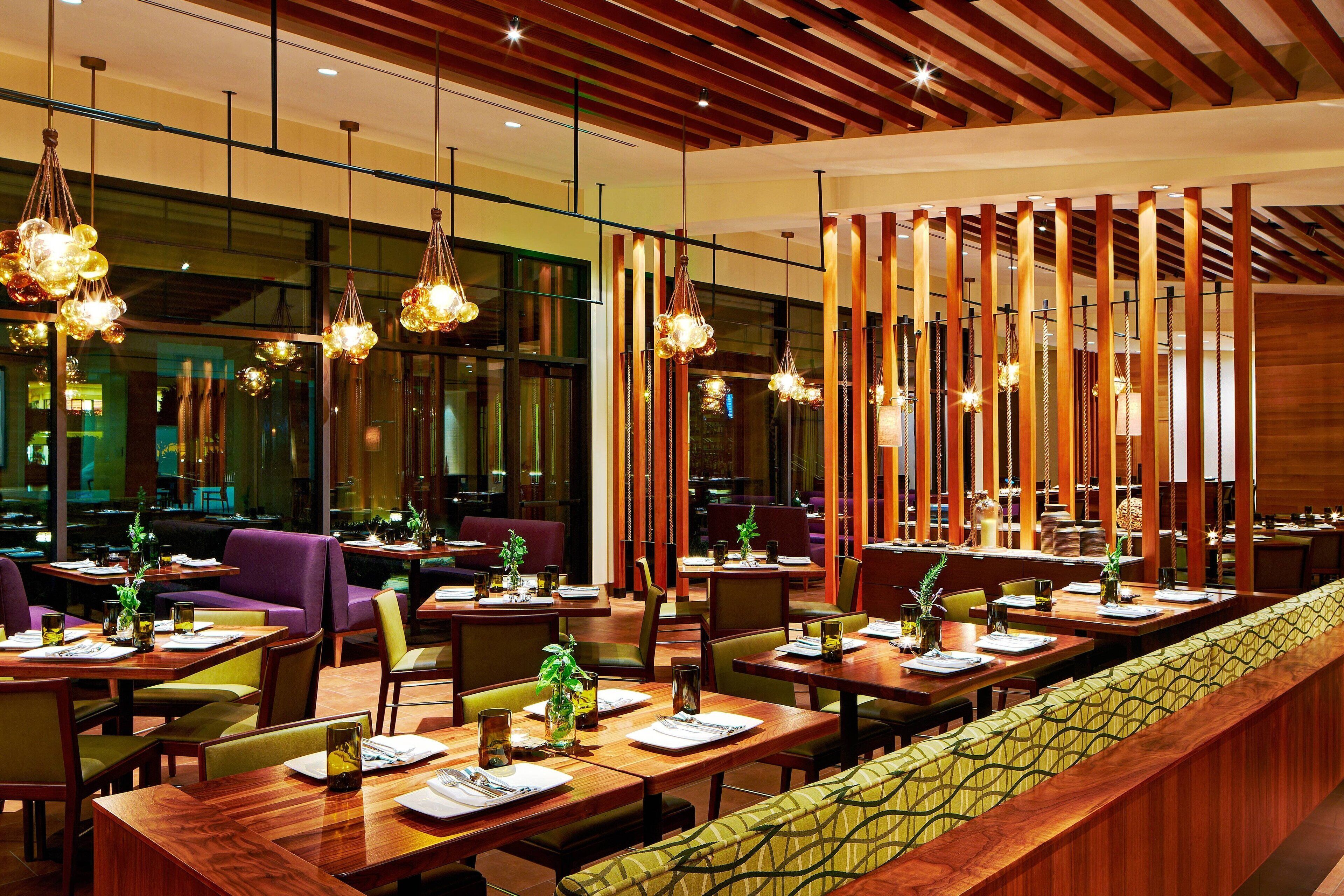 Un restaurante moderno con mesas de madera, asientos con cojines verdes e iluminación colgante.