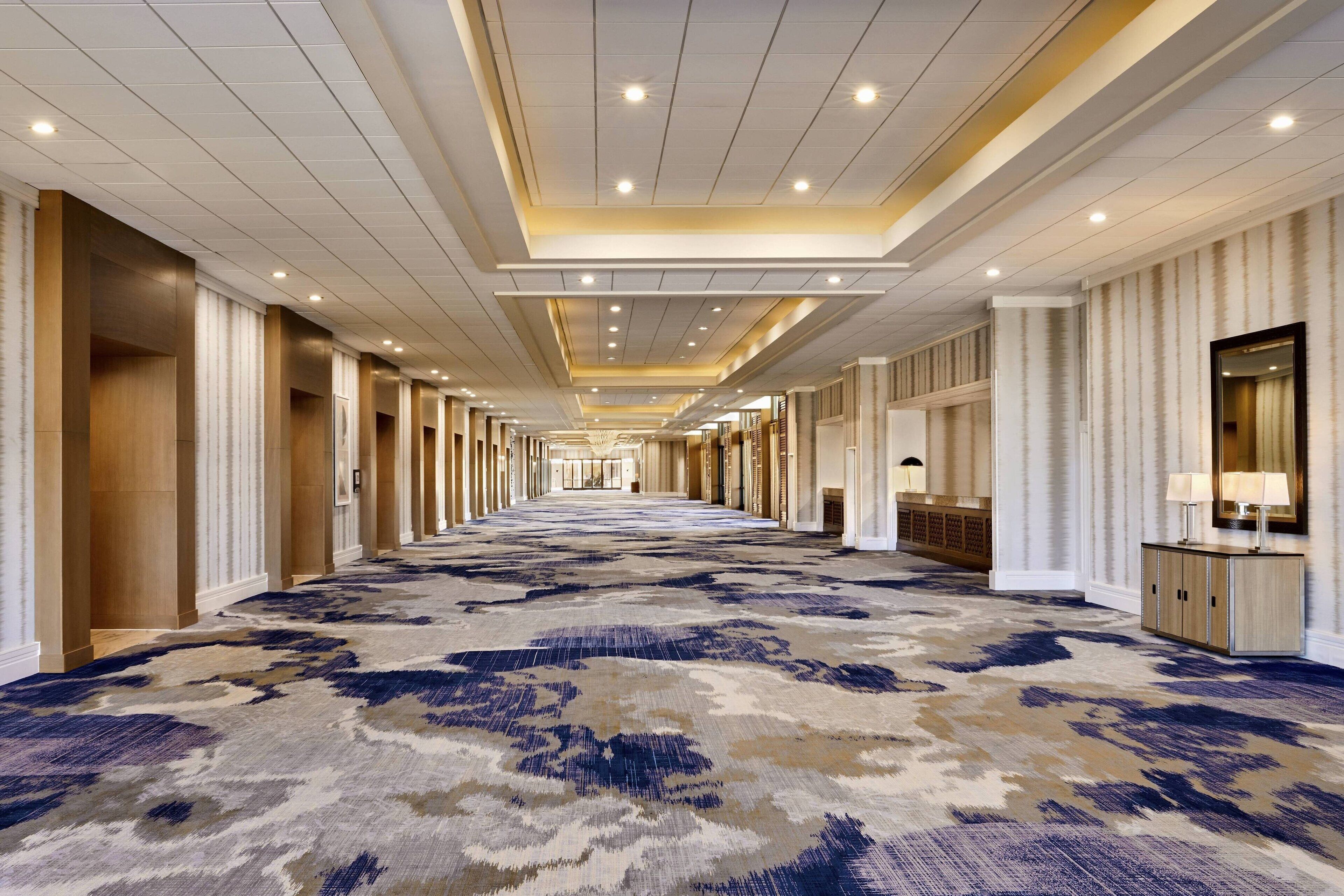 A long, narrow hotel corridor with a patterned carpet, recessed lighting, and a symmetrical design with columns and wall-mounted lamps.
