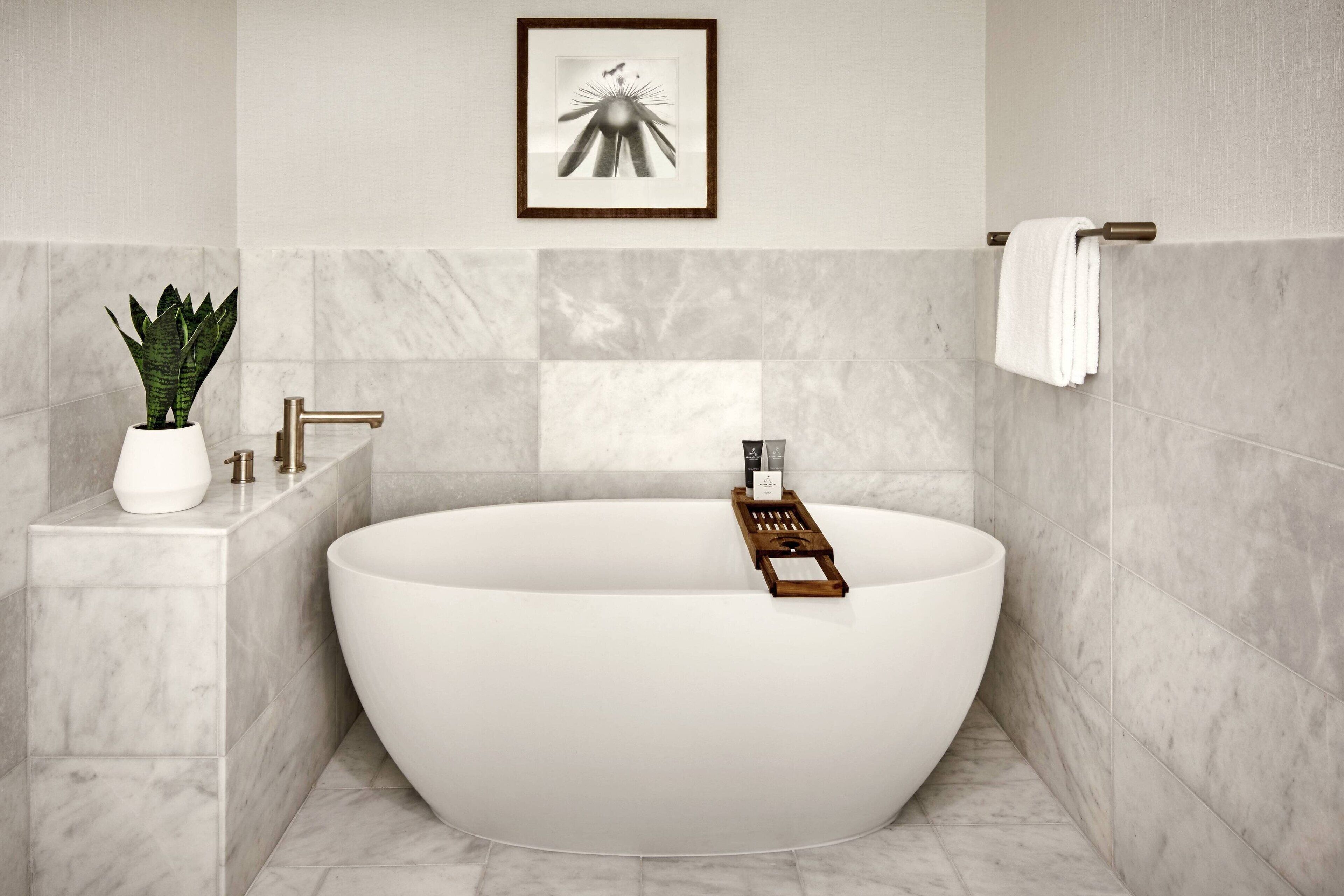 A modern bathroom with a freestanding bathtub, a potted plant, a towel rack, and a framed artwork on the wall.