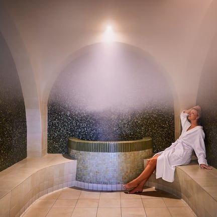 A person relaxing in a spa setting with a mosaic wall and a central hot tub.