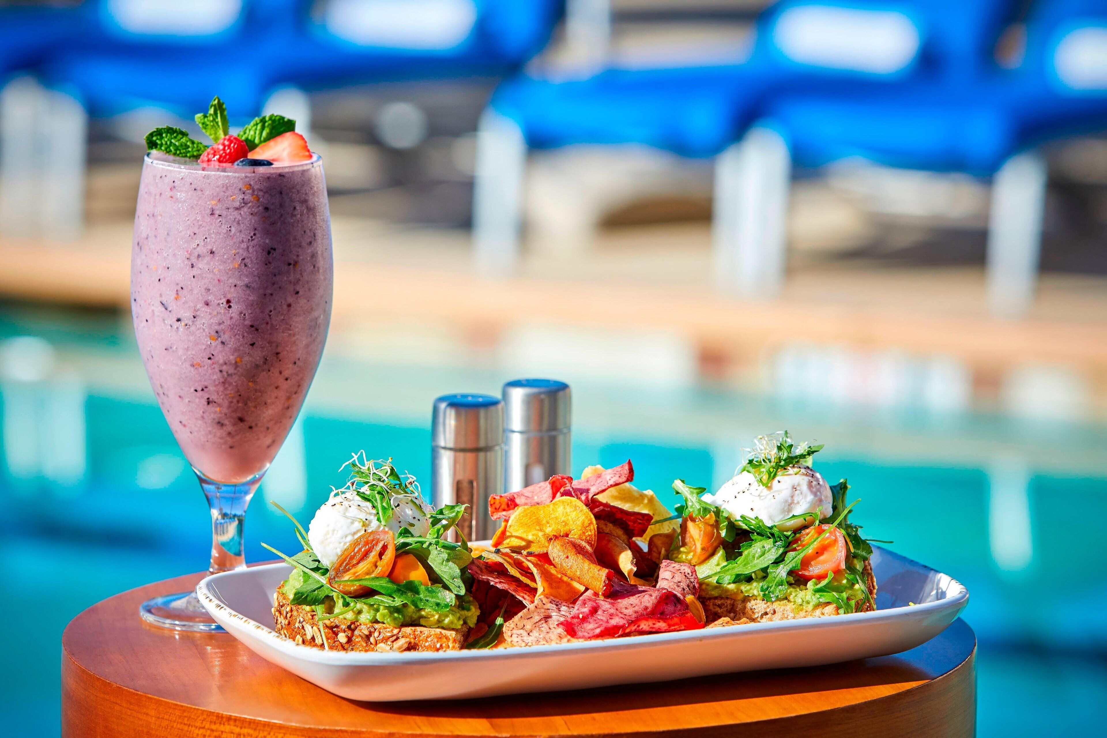 Un vaso de smoothie con una fresa en el borde, un plato de comida con verduras, tomates y un huevo pochado, y un ambiente junto a la piscina con sillas azules y una mesa de madera.