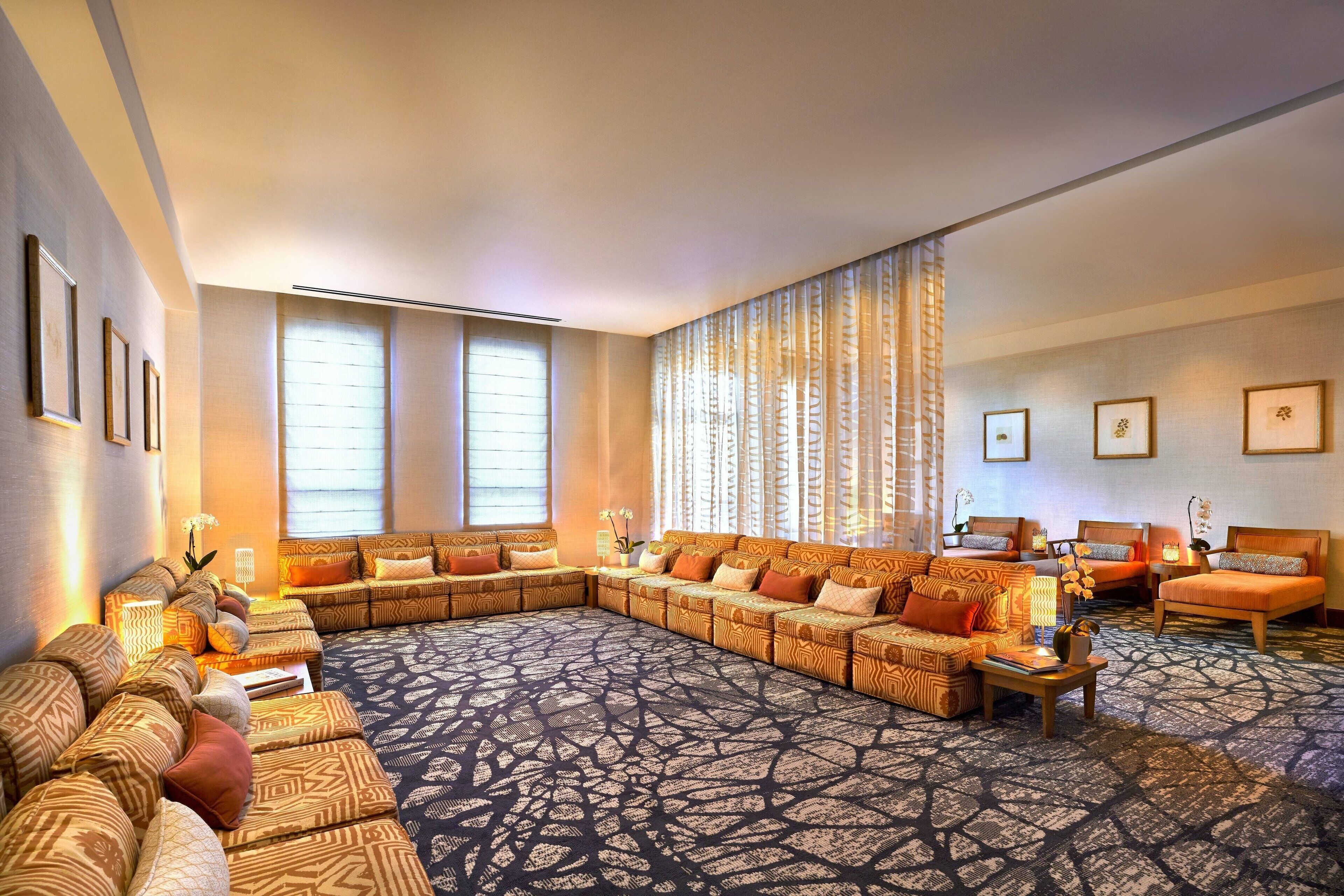 A spacious living room with a large sectional sofa, a separate sofa, and a couple of armchairs. There are multiple windows providing natural light, and the room features a patterned carpet.