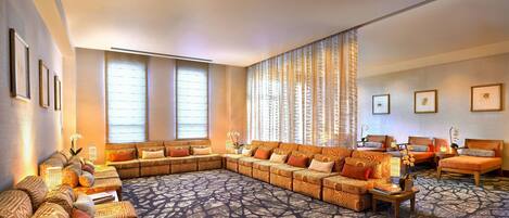 A spacious living room with a large sectional sofa, a separate sofa, and a couple of armchairs. There are multiple windows providing natural light, and the room features a patterned carpet.