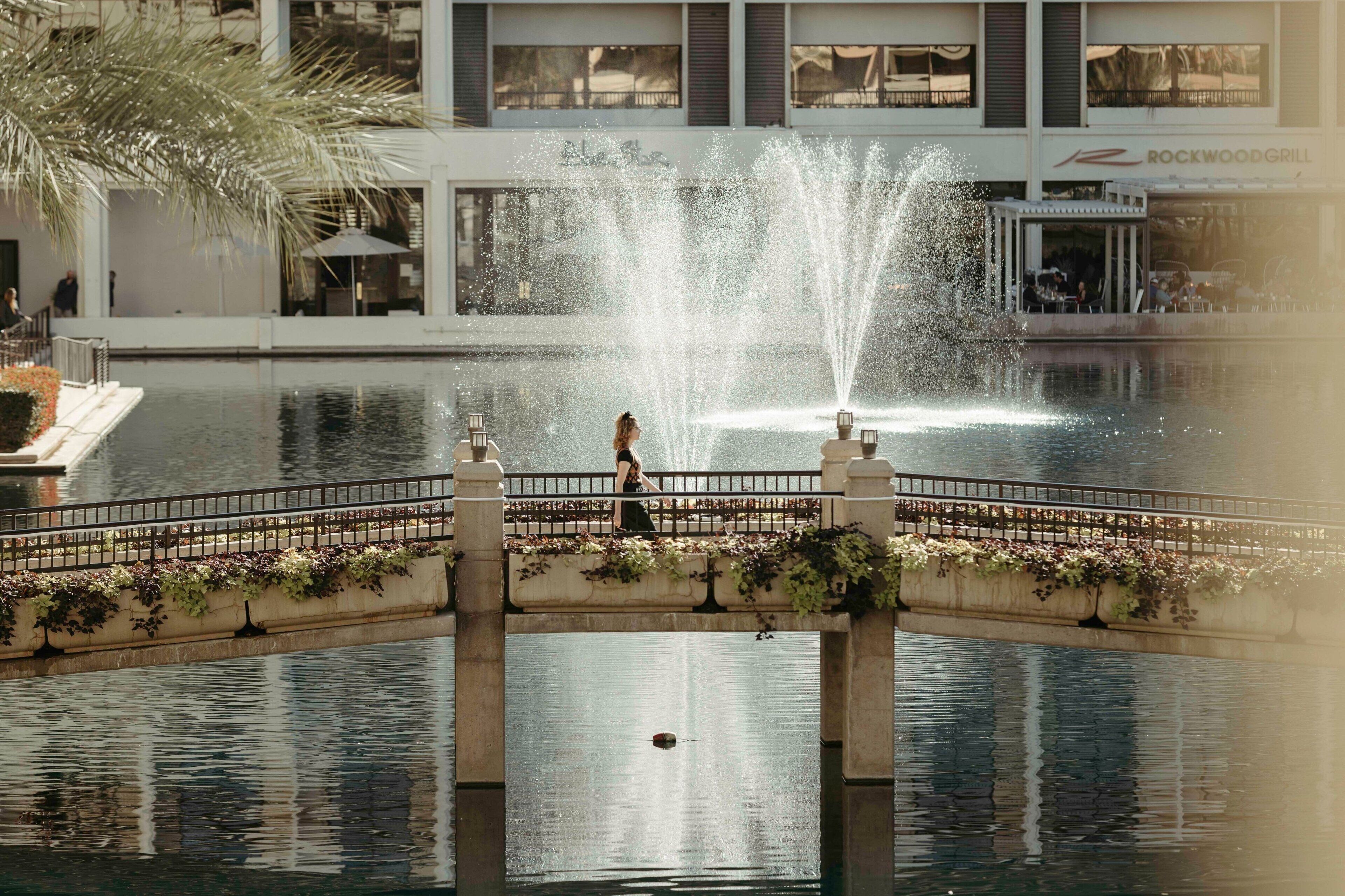 A bridge with a fountain in the middle, a pool, and a building with a restaurant in the background.