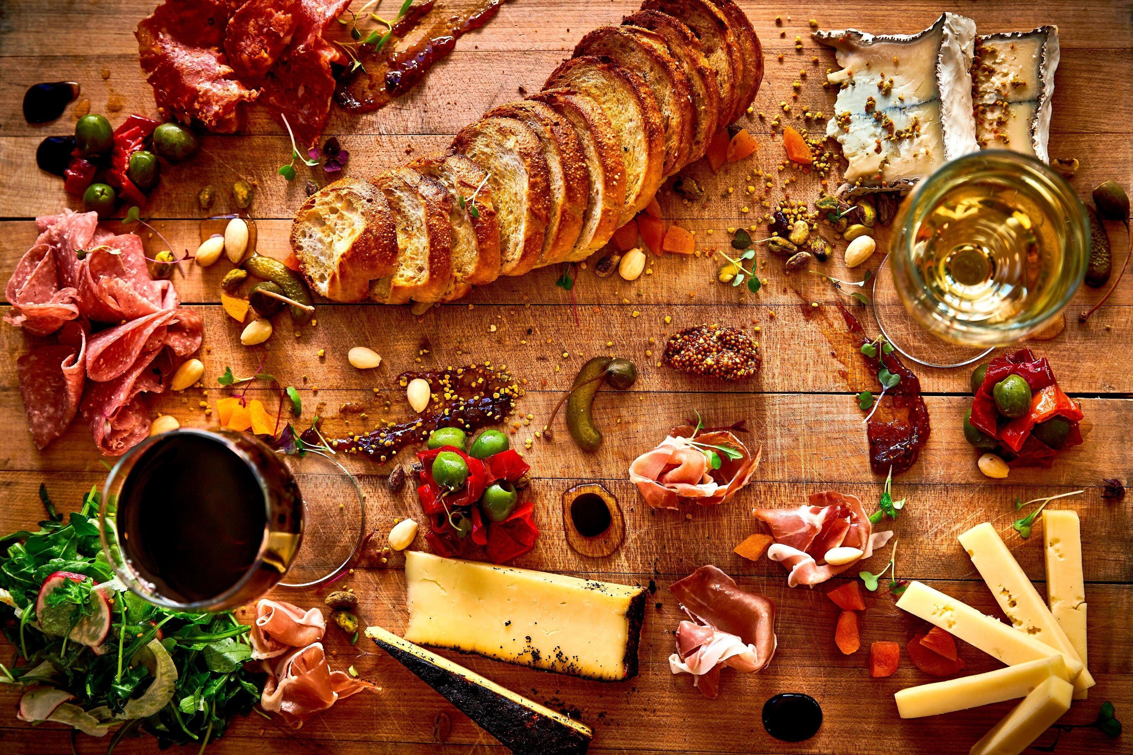 A wooden table with various food items including sliced bread, cheese, cured meats, olives, and nuts.