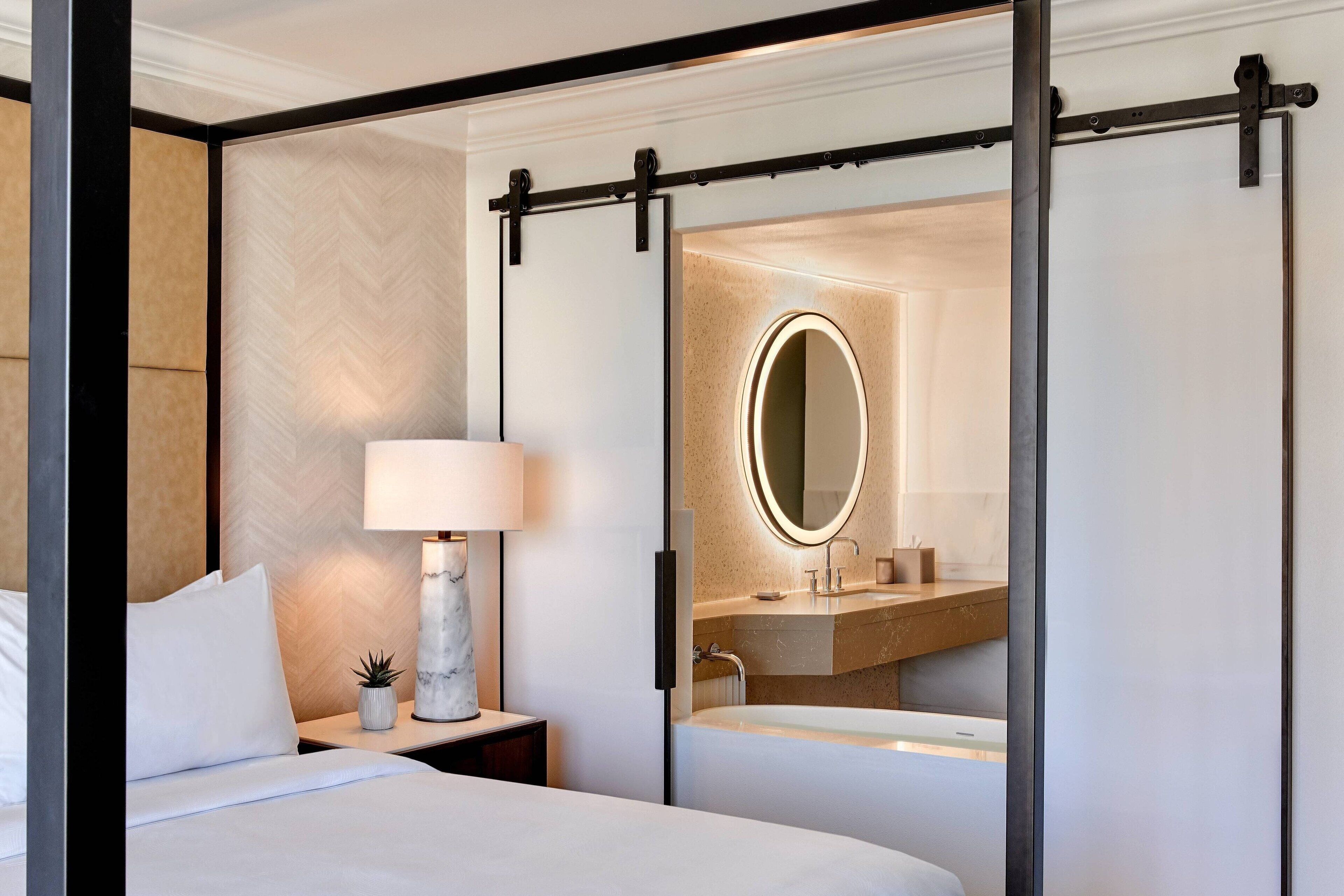 A modern hotel room with a bed, a nightstand with a lamp, a small plant, and a bathroom with a sink and mirror.