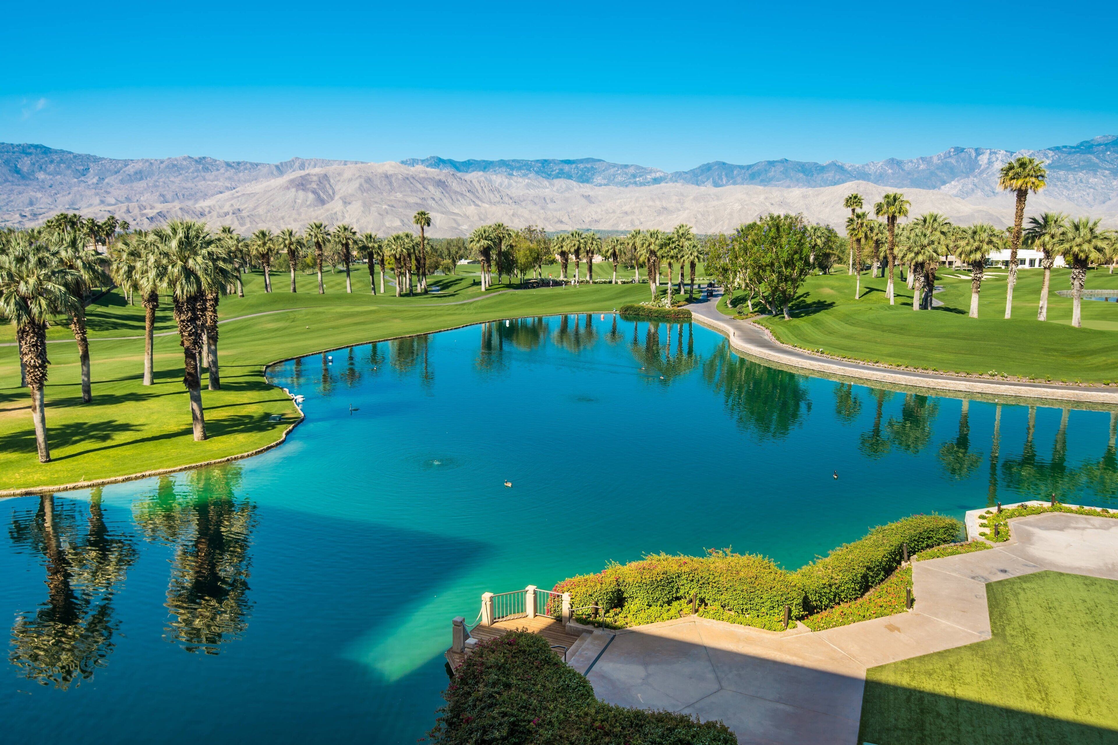 A golf course with a large pond, palm trees, and mountains in the background.