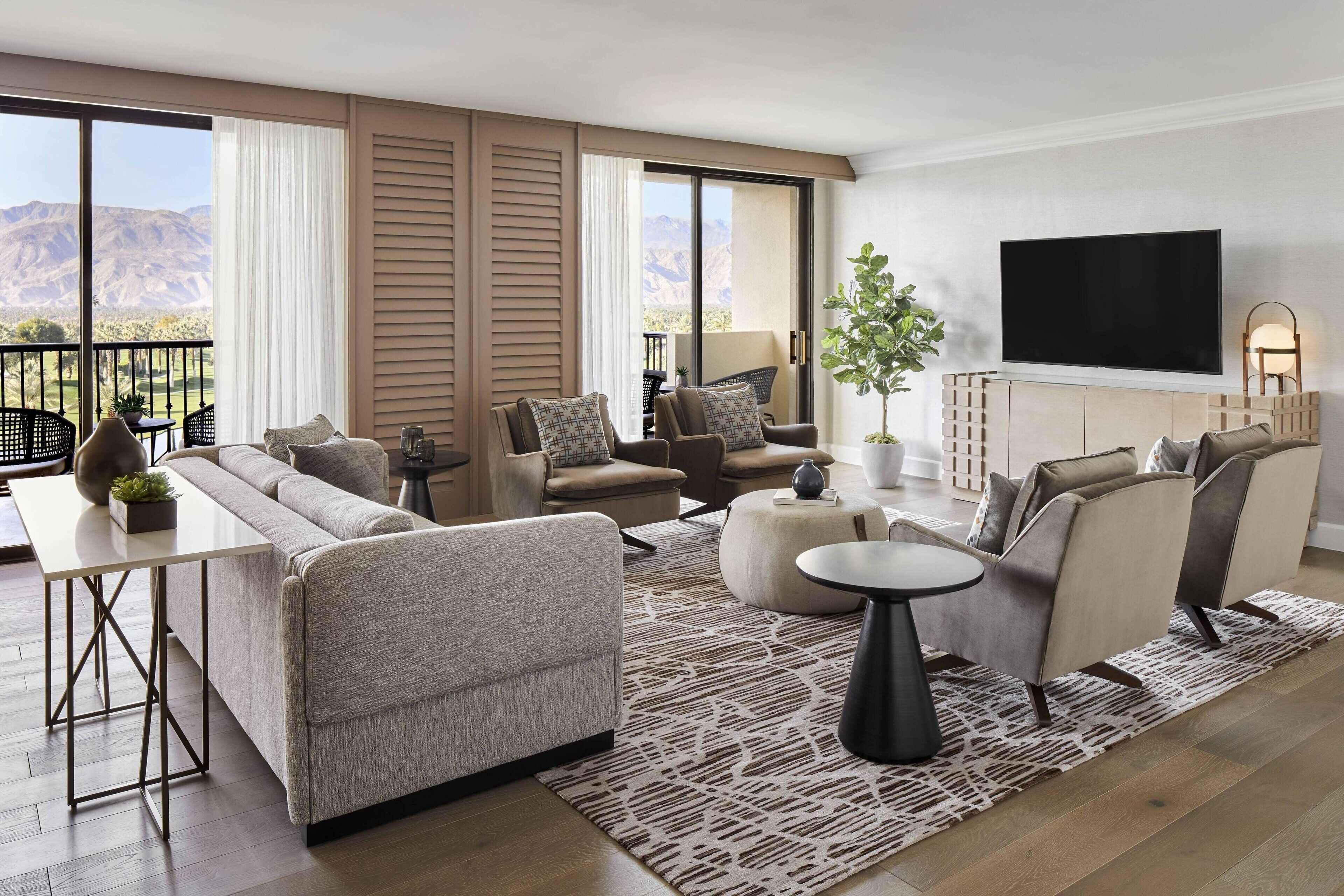Un salon moderne avec un canapé, des fauteuils, une table basse et un téléviseur. Il y a un balcon avec vue sur la montagne.