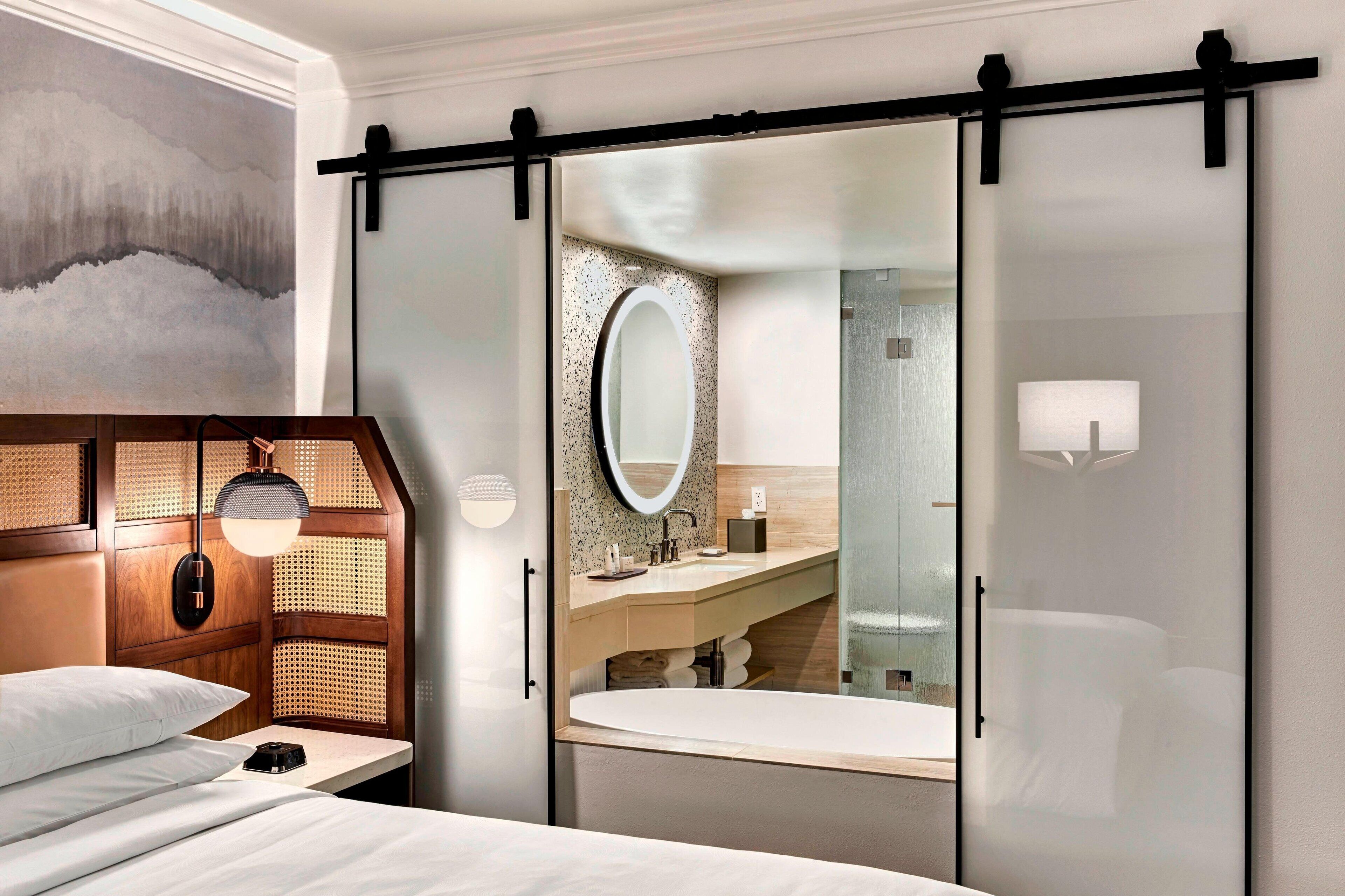 Et moderne hotelværelse med seng, træhovedgærde, et stort spejl, badeværelse med vask og toilet samt et skab med skydedøre.