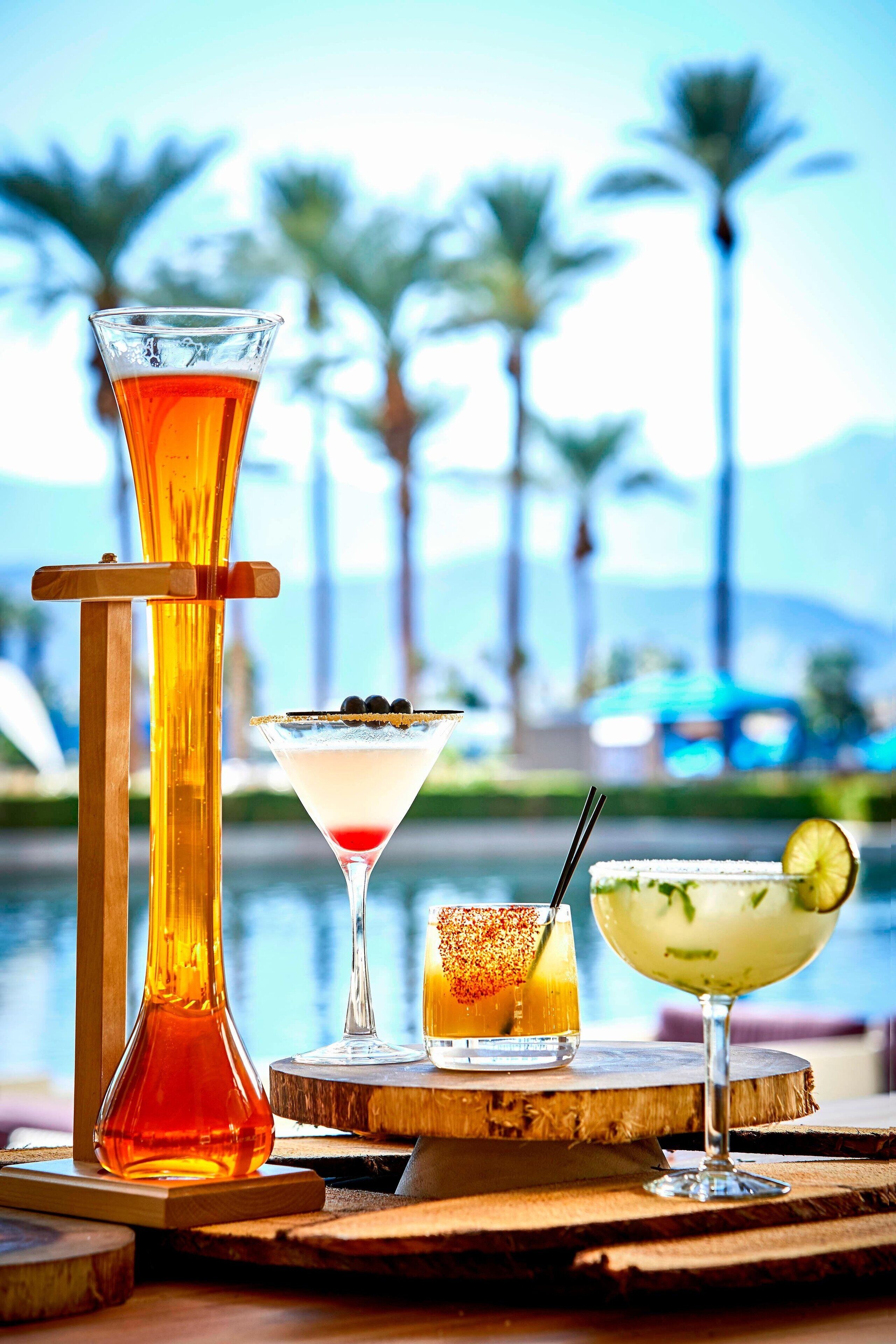 A variety of drinks on a wooden table with a pool and palm trees in the background.