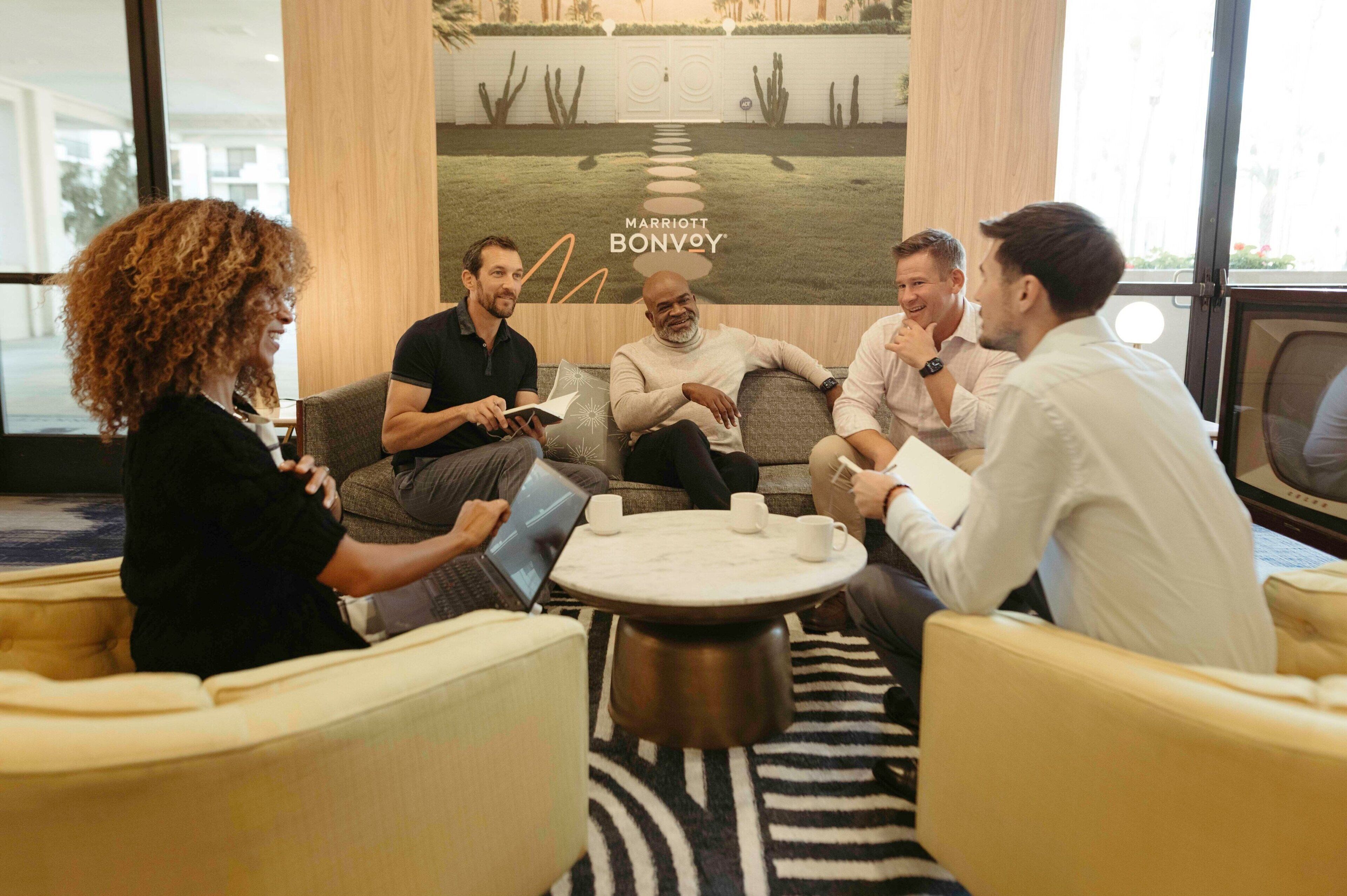 A group of people in a modern lounge area with a large poster of a desert landscape, a round coffee table, and comfortable seating.