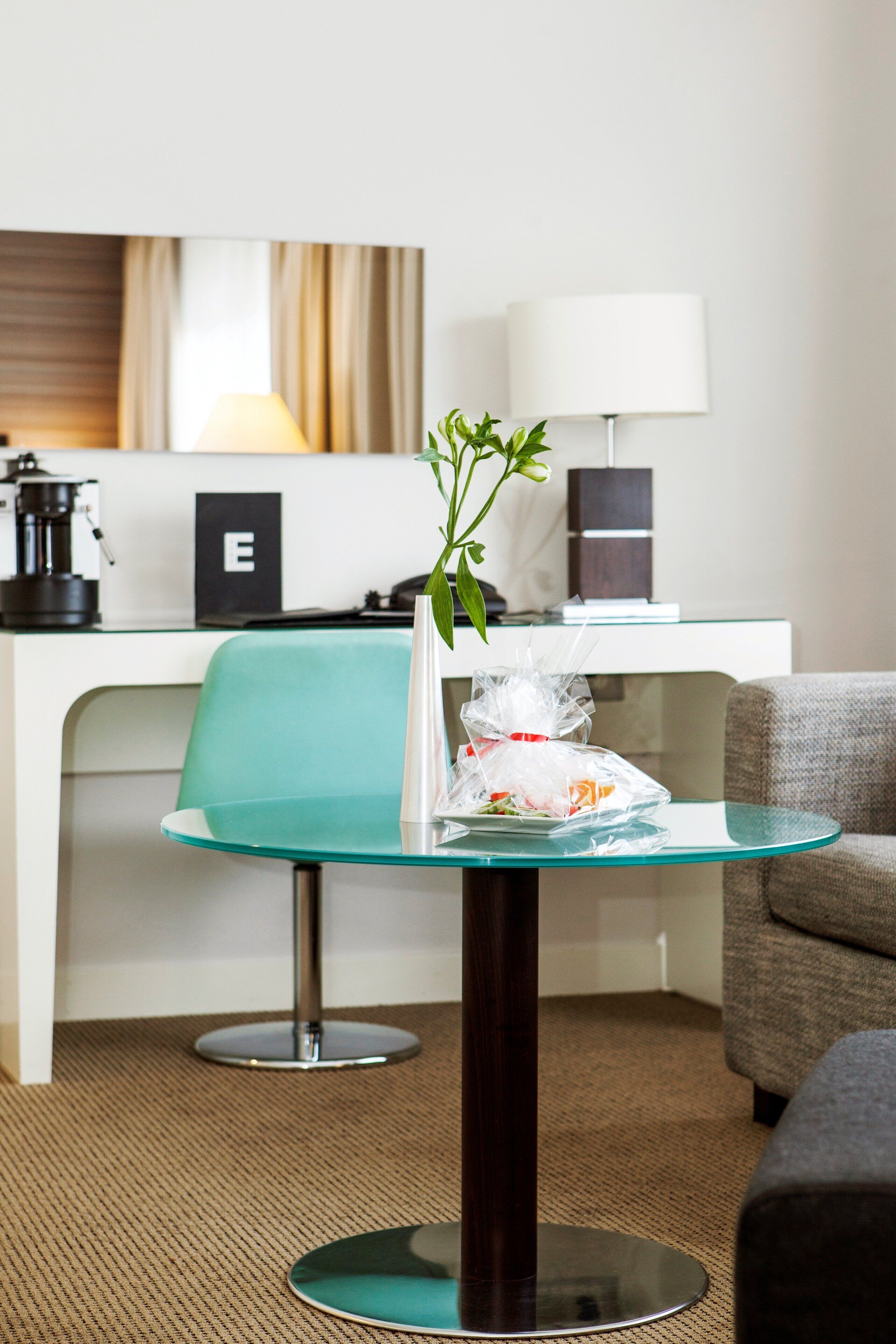 A hotel room with a glass table, a green chair, a sofa, a lamp, a coffee maker, and a plant.