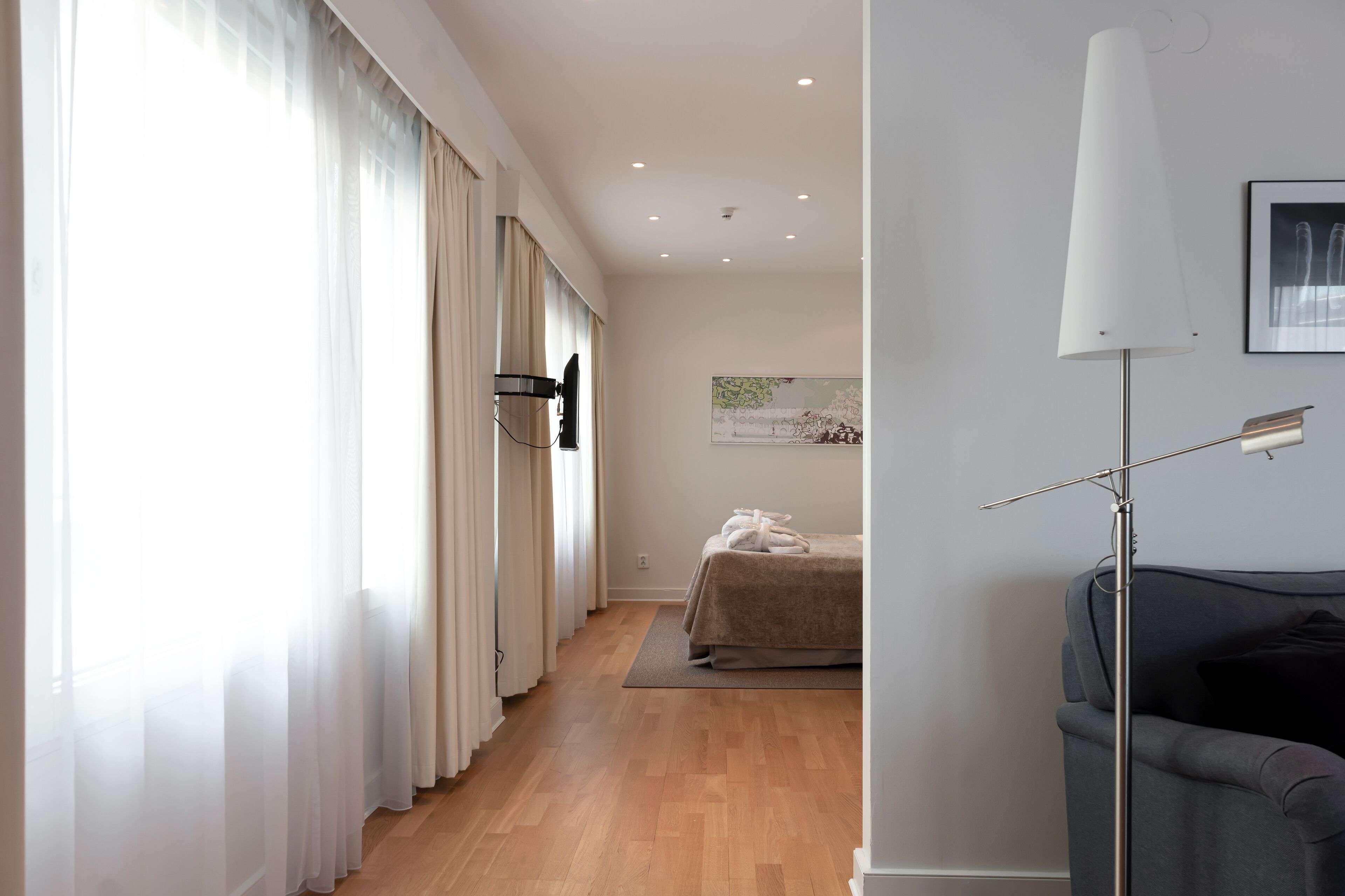 Una habitación de hotel moderna con piso de madera, una cama con cubierta color beige, un ventanal con cortinas translúcidas, una lámpara de pie y un cuadro en la pared.