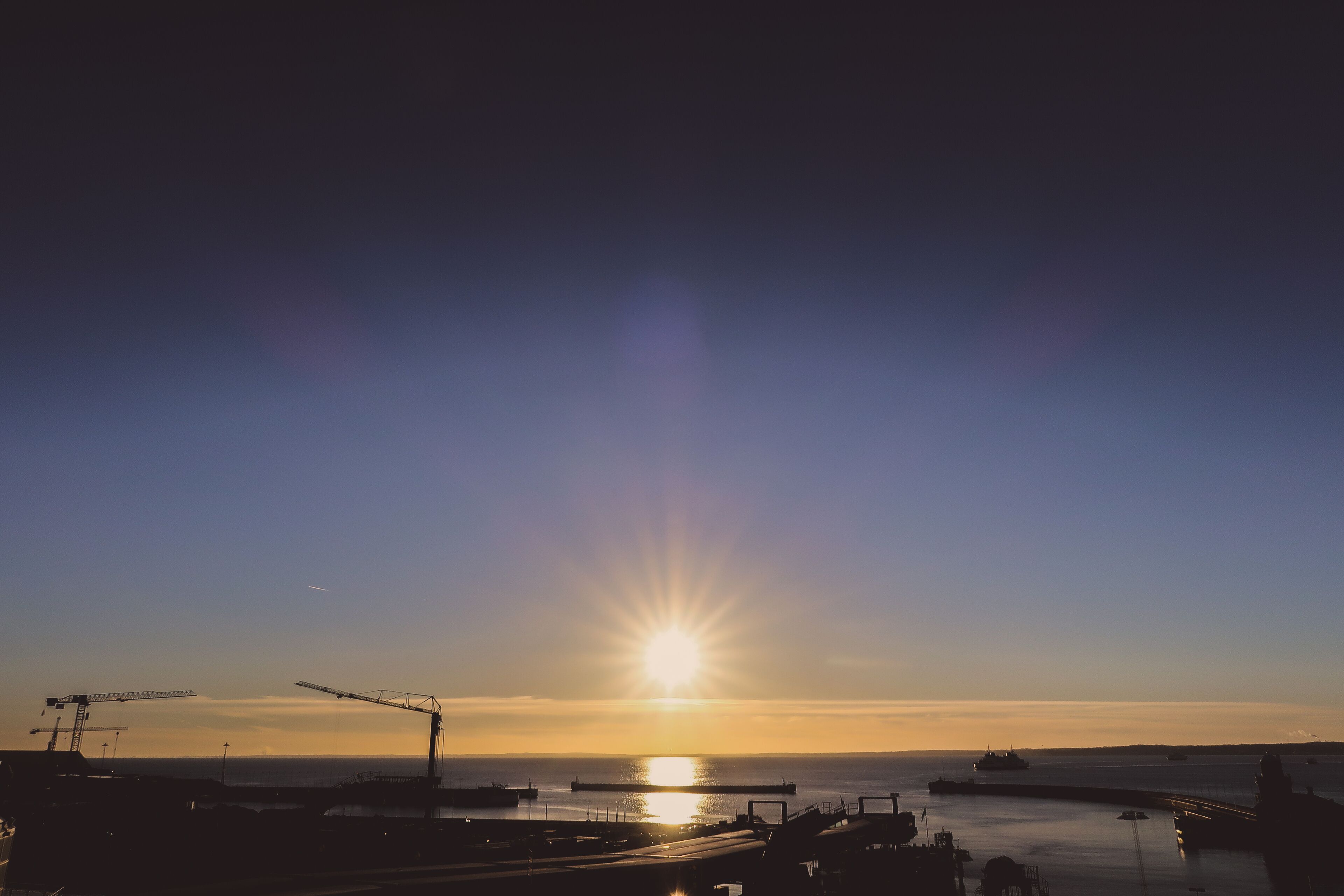 A sunset over a harbor with cranes and boats.