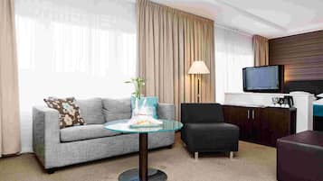 Una habitación de hotel moderna con un sofá gris, un sillón negro, una mesa de centro de cristal, un televisor y un mueble de madera.