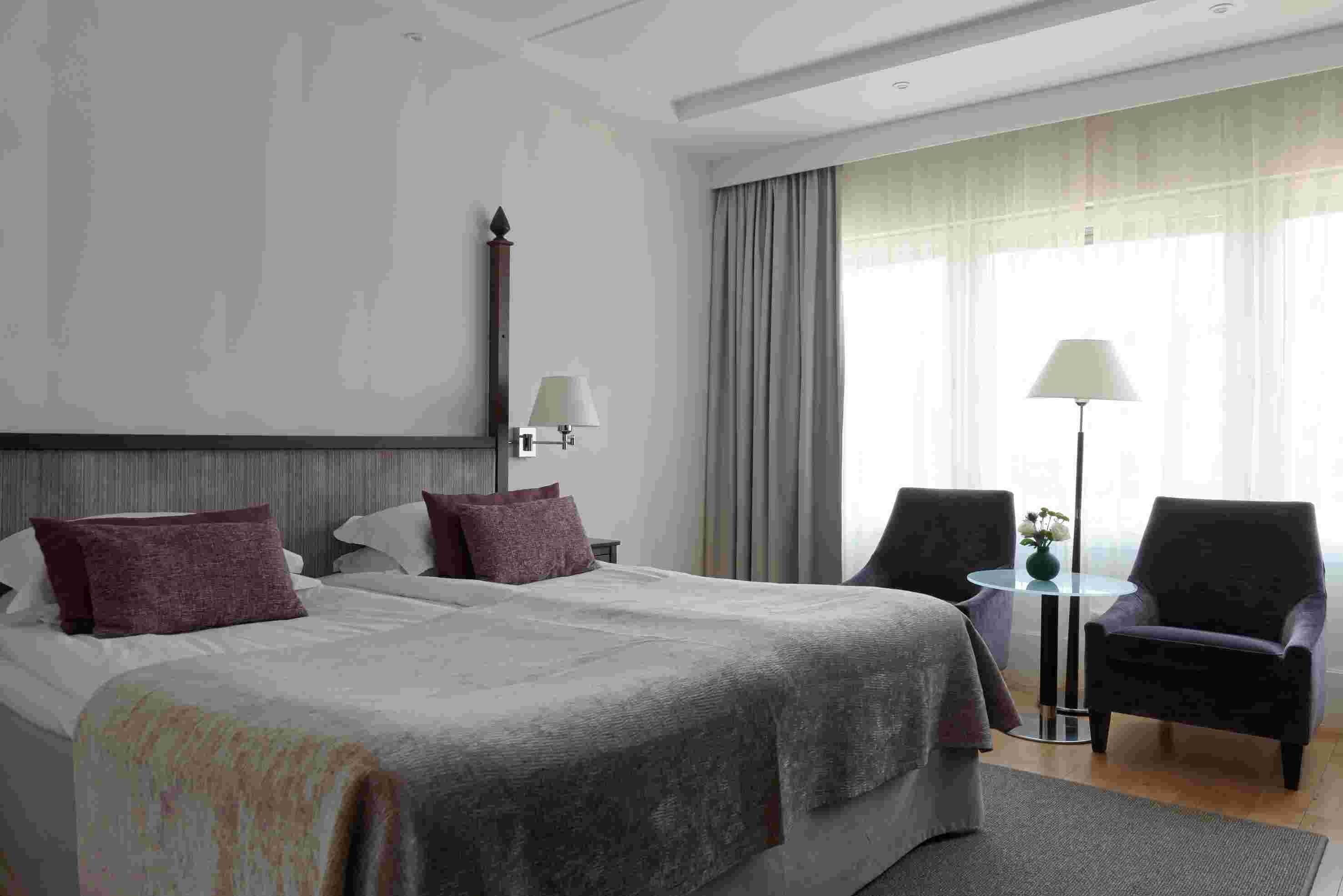 Ett hotellrum med en stor säng, två stolar, ett litet bord och en lampa.