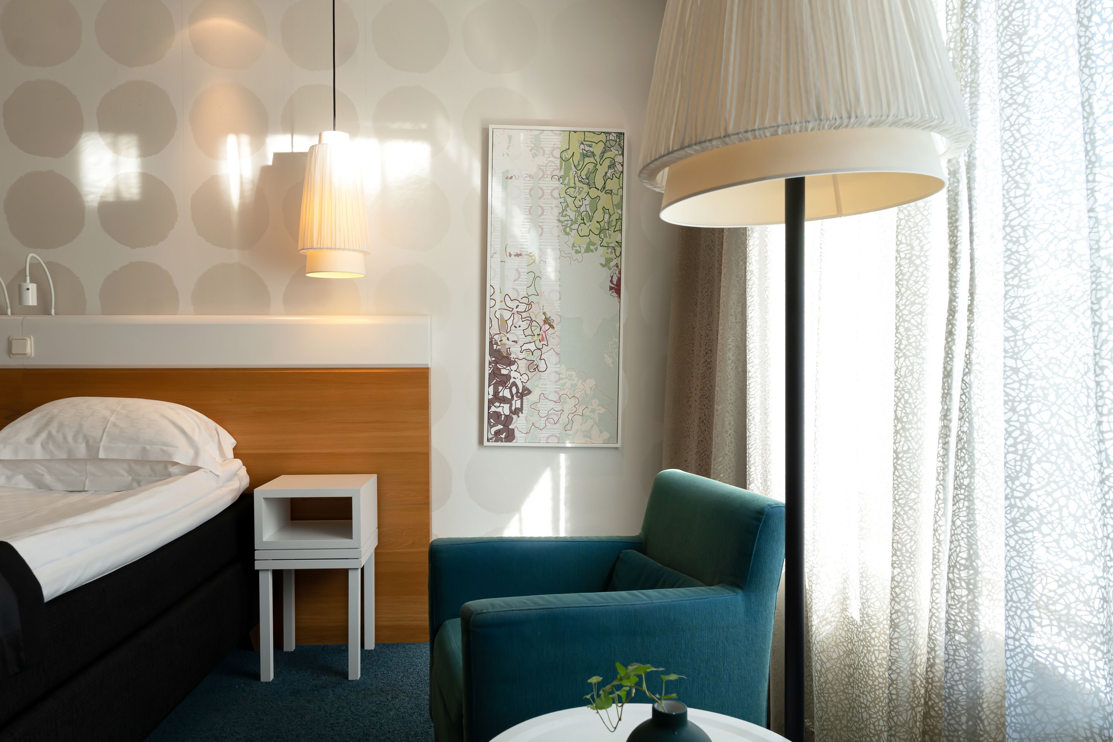 Quarto de hotel com cama, pequena mesa de cabeceira, poltrona azul, mesa redonda com um vaso, luminária de parede e uma janela grande com cortinas translúcidas.