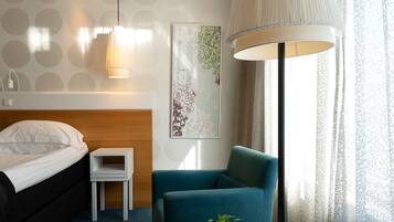 Quarto de hotel com cama, pequena mesa de cabeceira, poltrona azul, mesa redonda com um vaso, luminária de parede e uma janela grande com cortinas translúcidas.